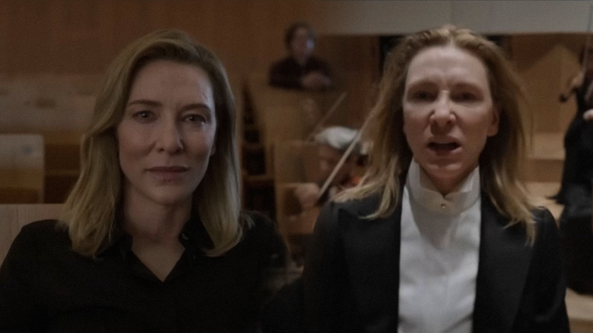 Actress Cate Blanchett in a black suit glares menacingly at the camera against a backdrop of violinists