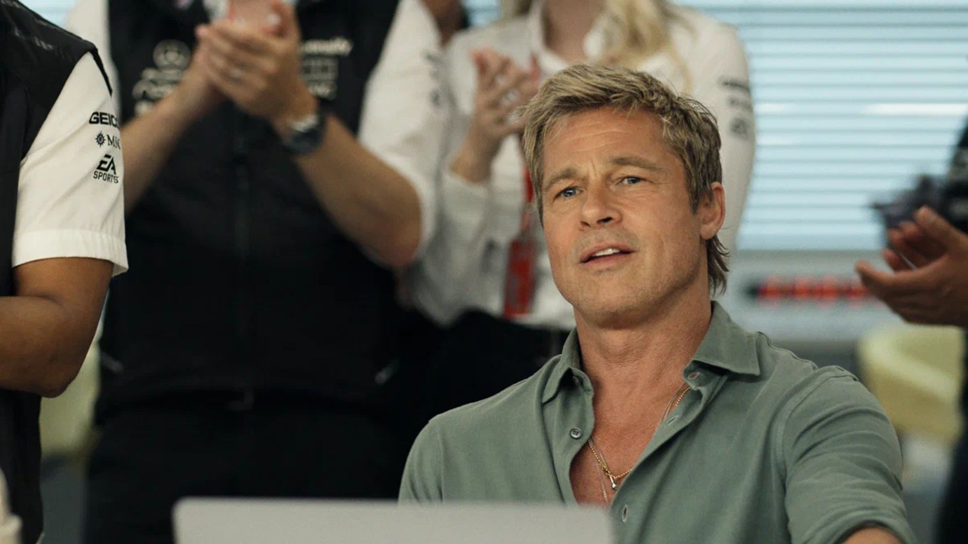 Brad Pitt in an unbuttoned shirt sits in front of a laptop with applauding people in the background