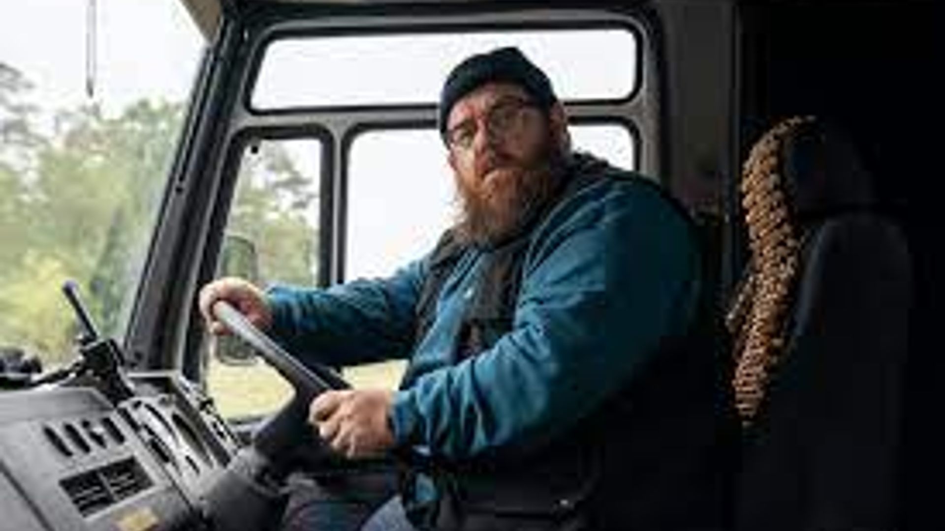 Nick Frost wearing glasses and a hat sits behind the wheel of a truck, looking at the camera