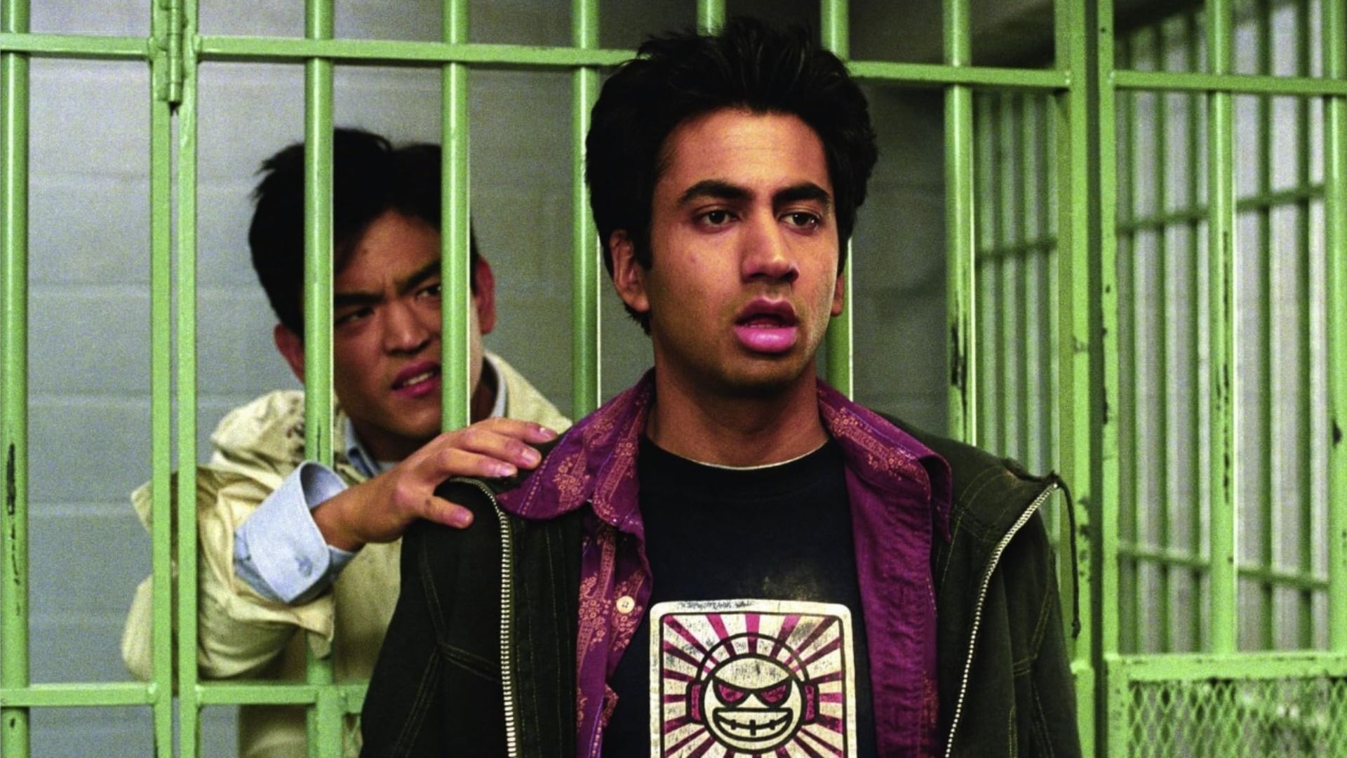 Actor John Cho stands behind green prison bars, actor Kal Penn stands outside and looks to the side