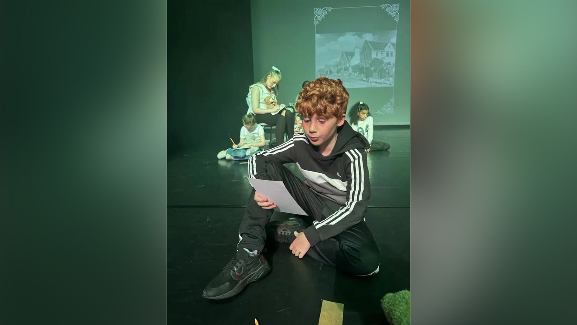 Alastair Stout in a tracksuit sitting on a theater stage reading from a sheet