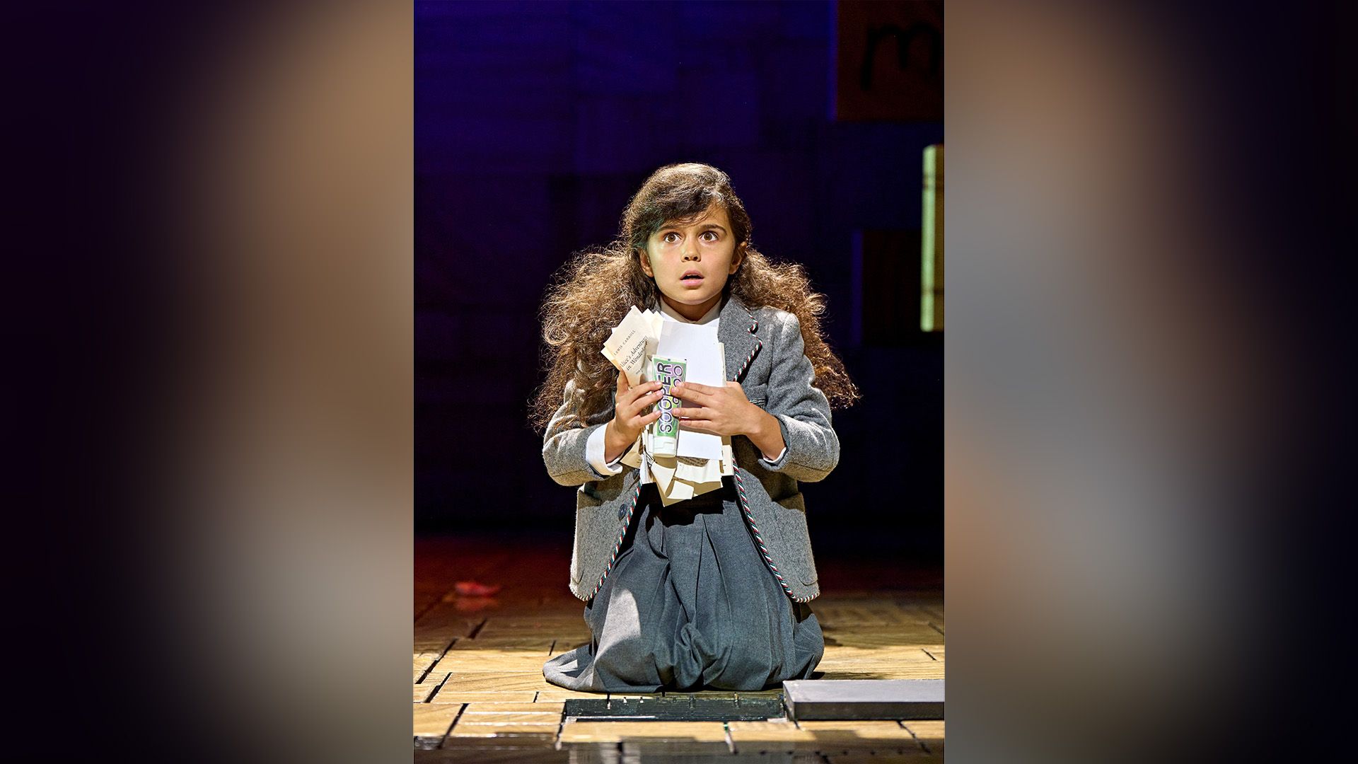 Arabella Stanton in a gray dress kneeling on a theater stage, holding crumpled sheets of paper in her hands