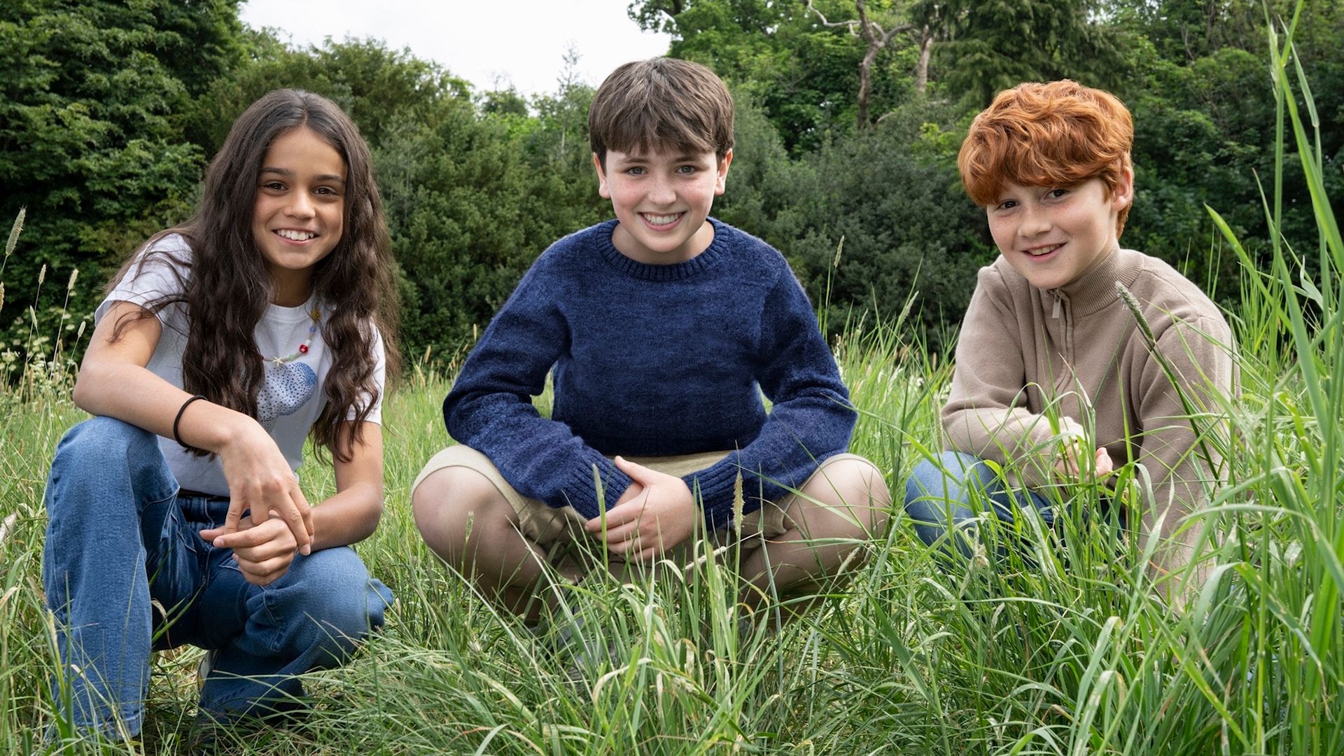 Young actors Arabella Stanton, Dominic McLaughlin, and Alastair Stout crouching in the grass and smiling