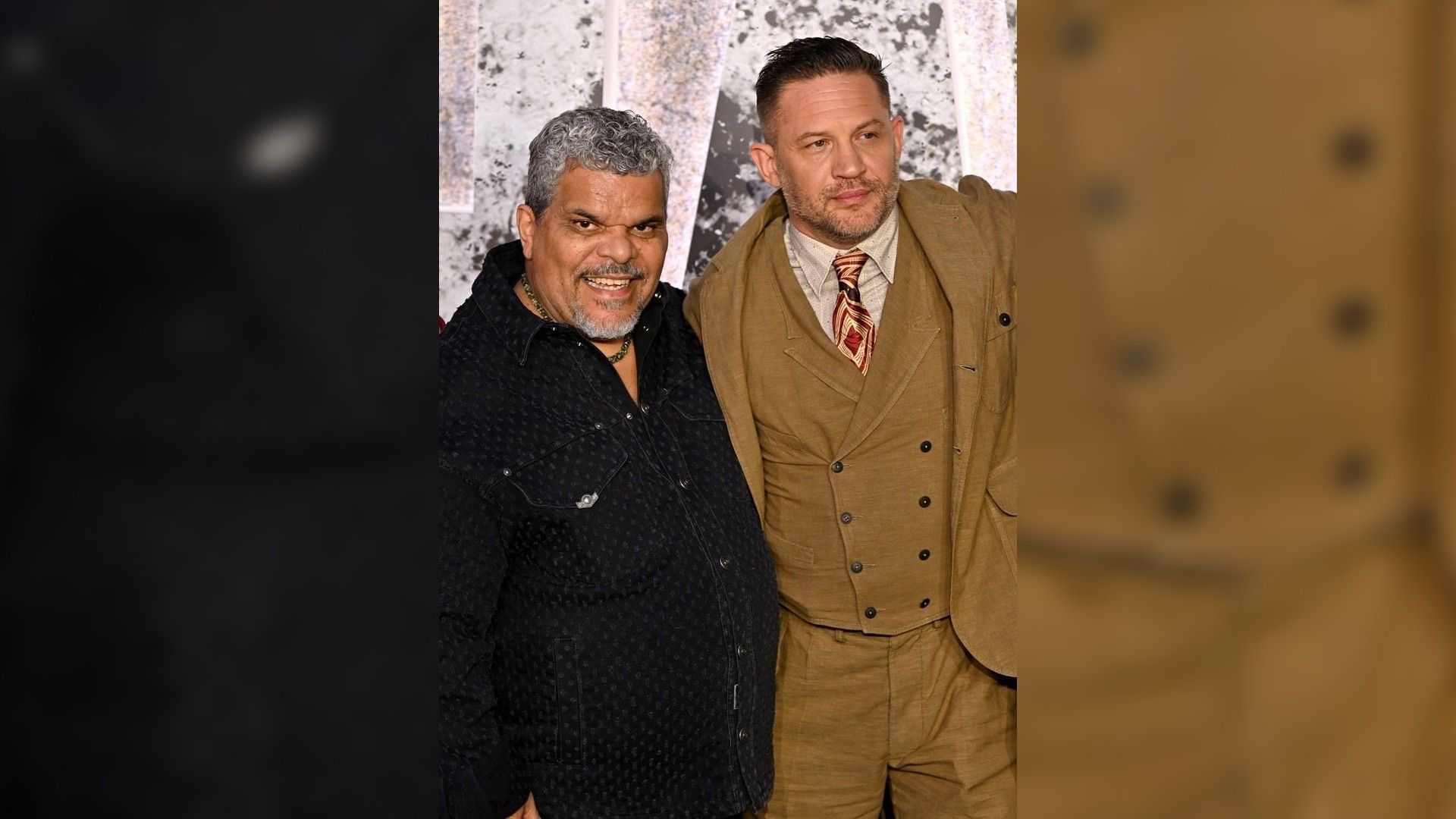 Luis Guzmán and Tom Hardy pose on the red carpet: one in a dark shirt, the other in a brown suit with a tie
