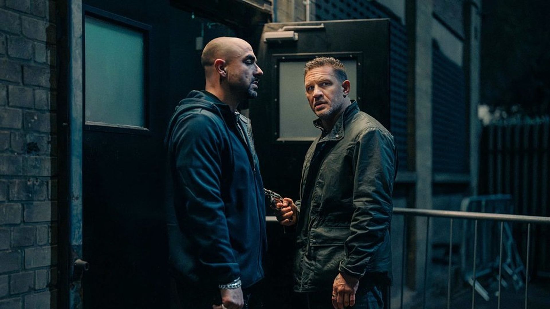 Tom Hardy and another man in dark clothing are talking by a black exit at night, one of them is holding a weapon