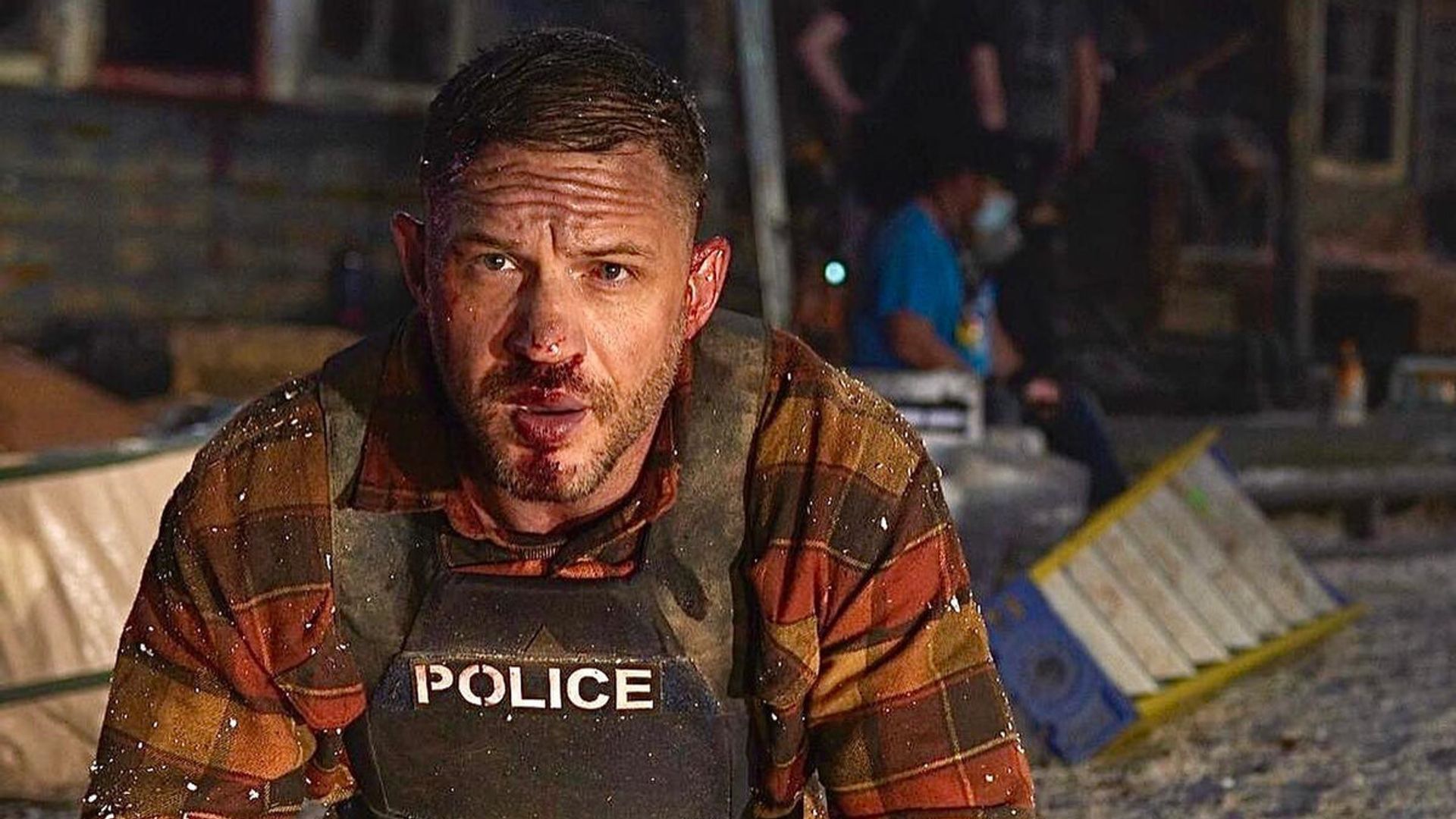 Tom Hardy in a bulletproof vest with a bruised face is kneeling at the scene of the incident