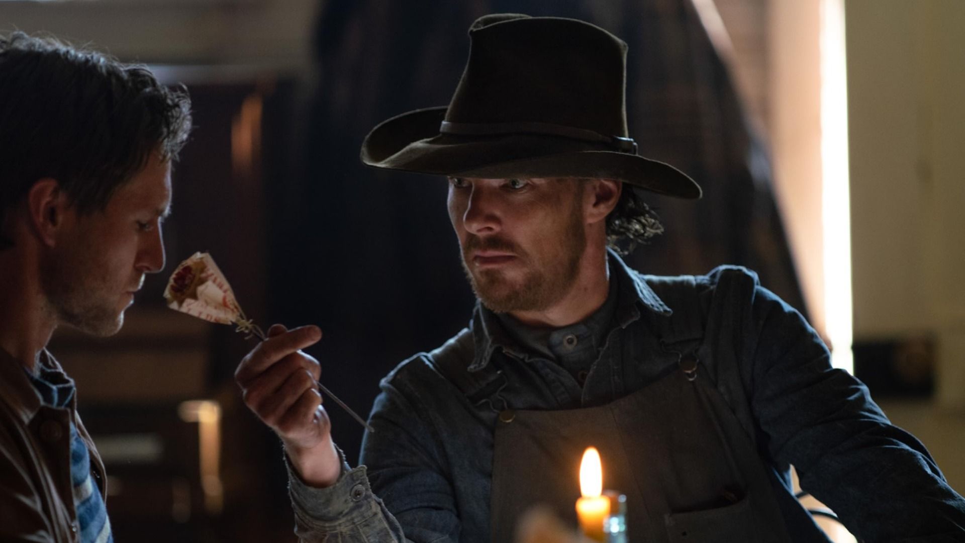A man in a hat brought a flower to another man's nose and is looking at him