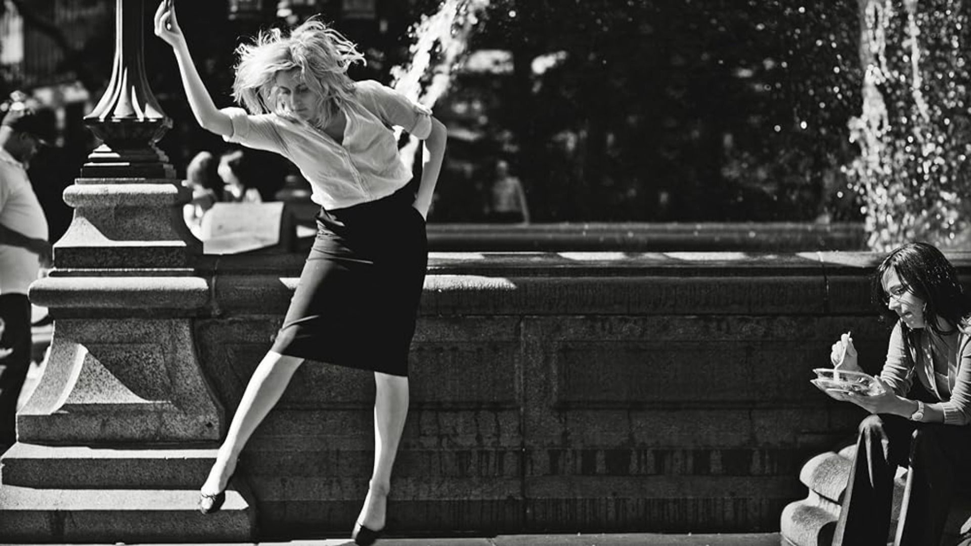 Actress Greta Gerwig dances in a white blouse and black skirt against the backdrop of a fountain