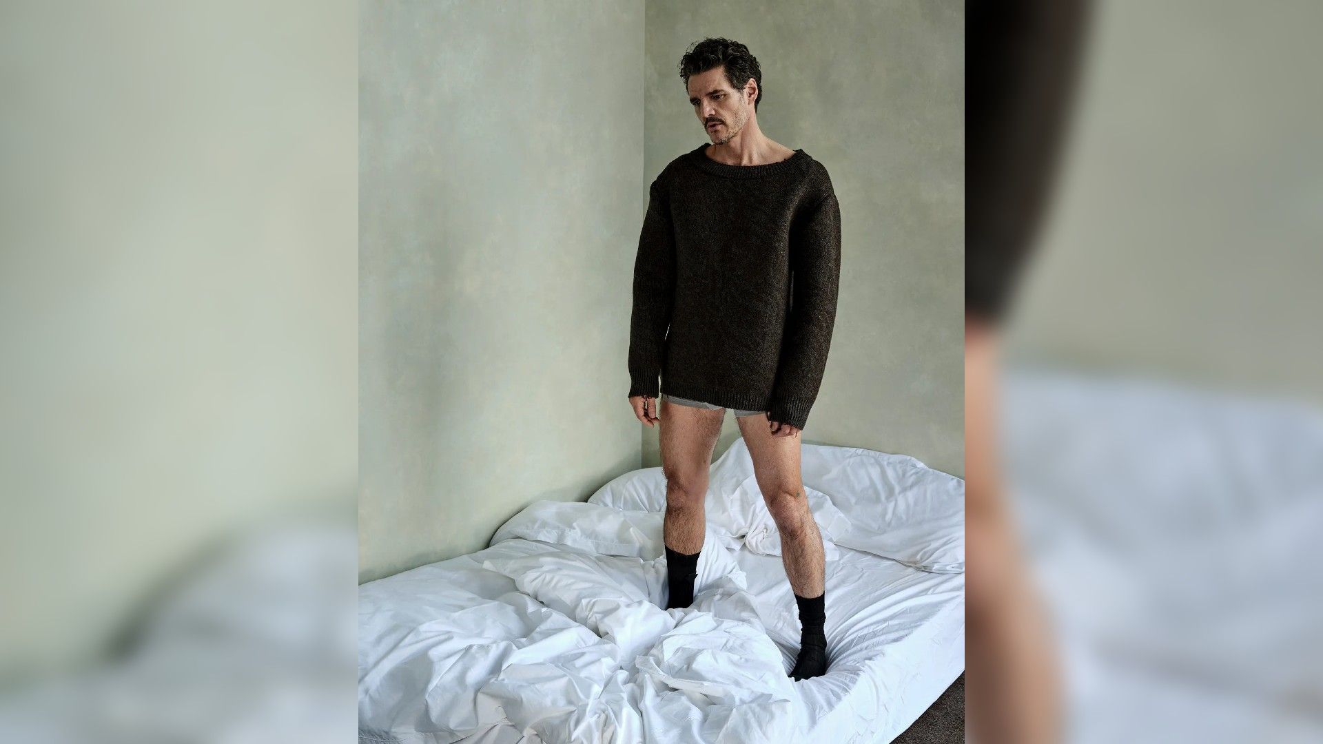 Pedro Pascal standing on a bed in a sweater and underwear