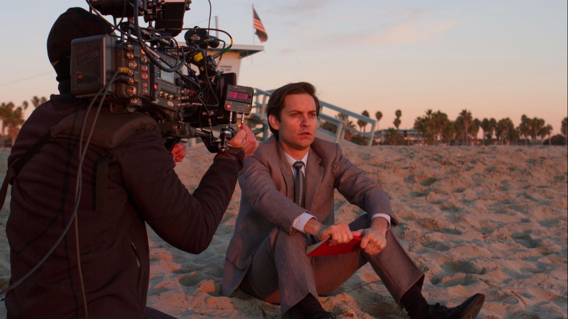 Actor Tobey Maguire in a gray suit sits on the sand