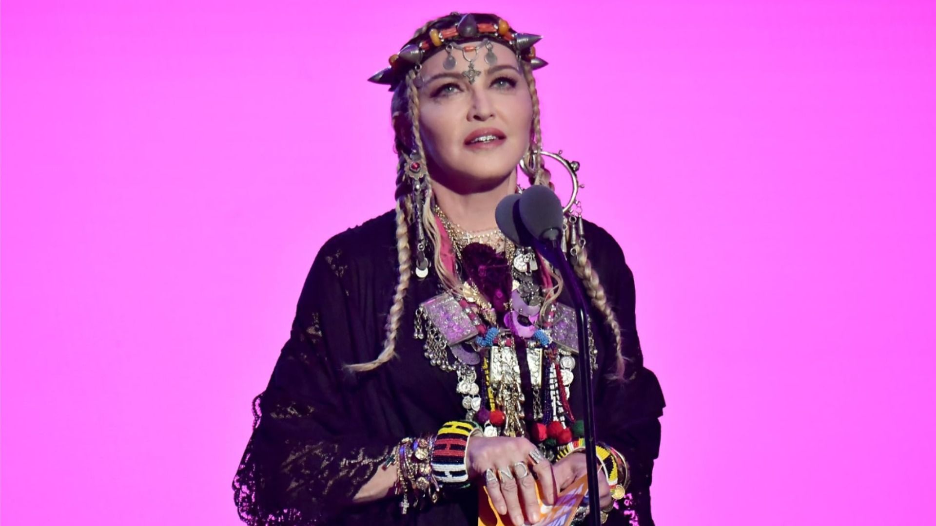 Singer Madonna stands in a black dress against a pink background and looks up