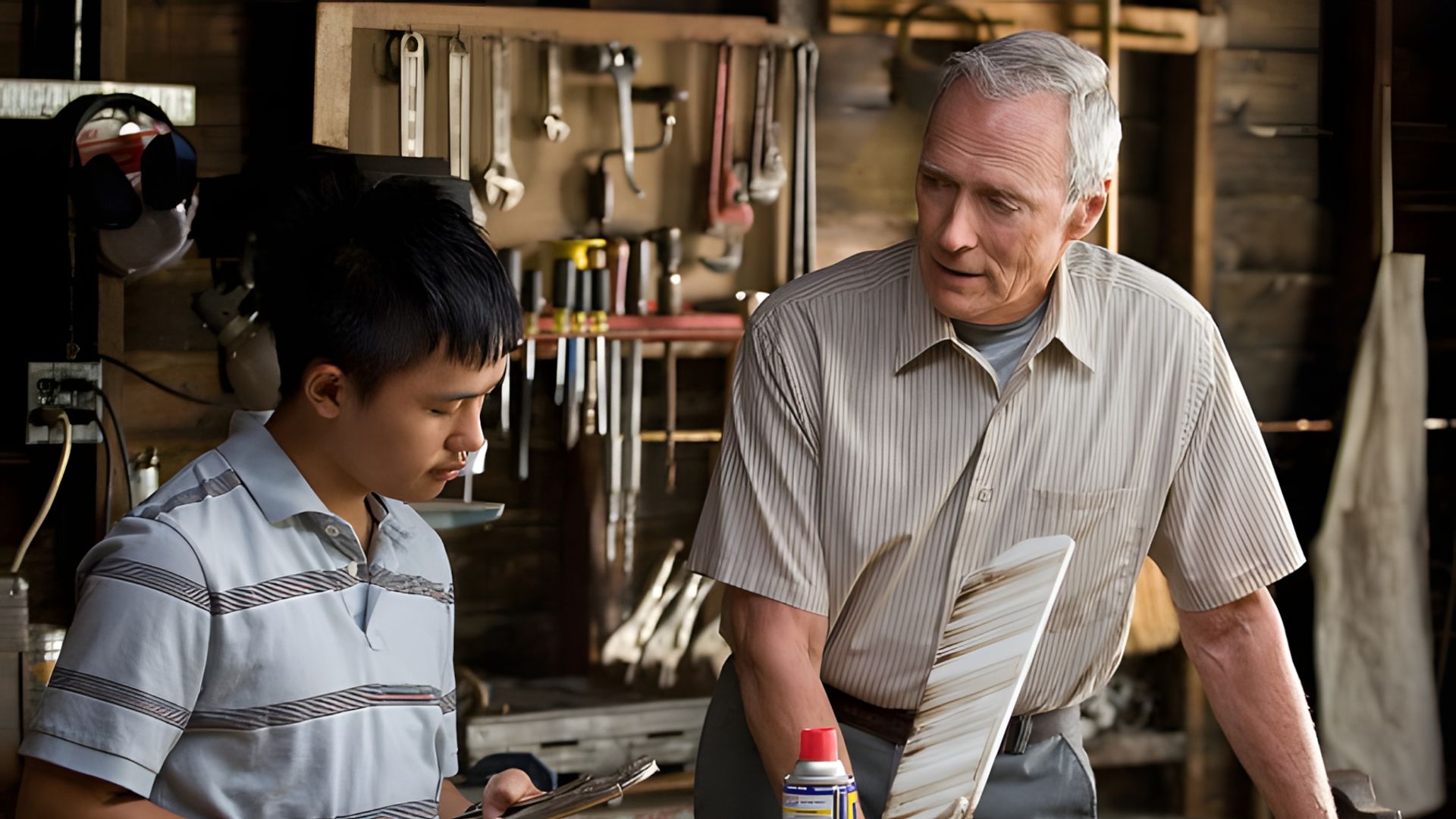 Clint Eastwood and a teenager work in a workshop, discussing a tool against the backdrop of a workbench with hanging tools