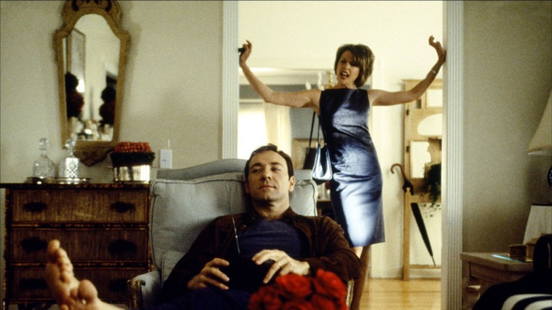 Kevin Spacey sits relaxed in a chair with a remote control while a woman in an evening dress behind him gesticulates emotionally in the doorway