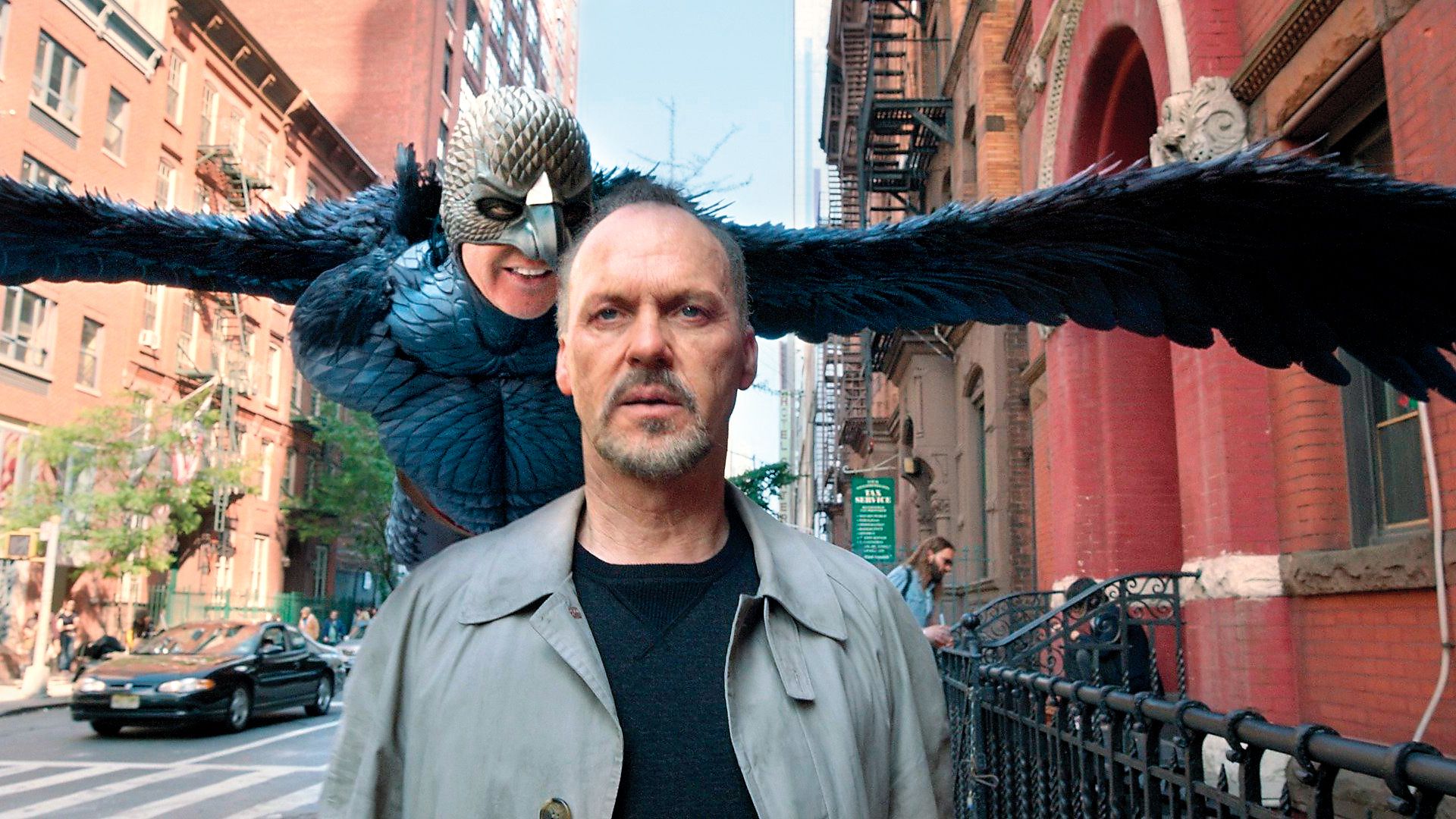 Michael Keaton walks down the street while behind him hovers a fantastical character with wings and a bird mask
