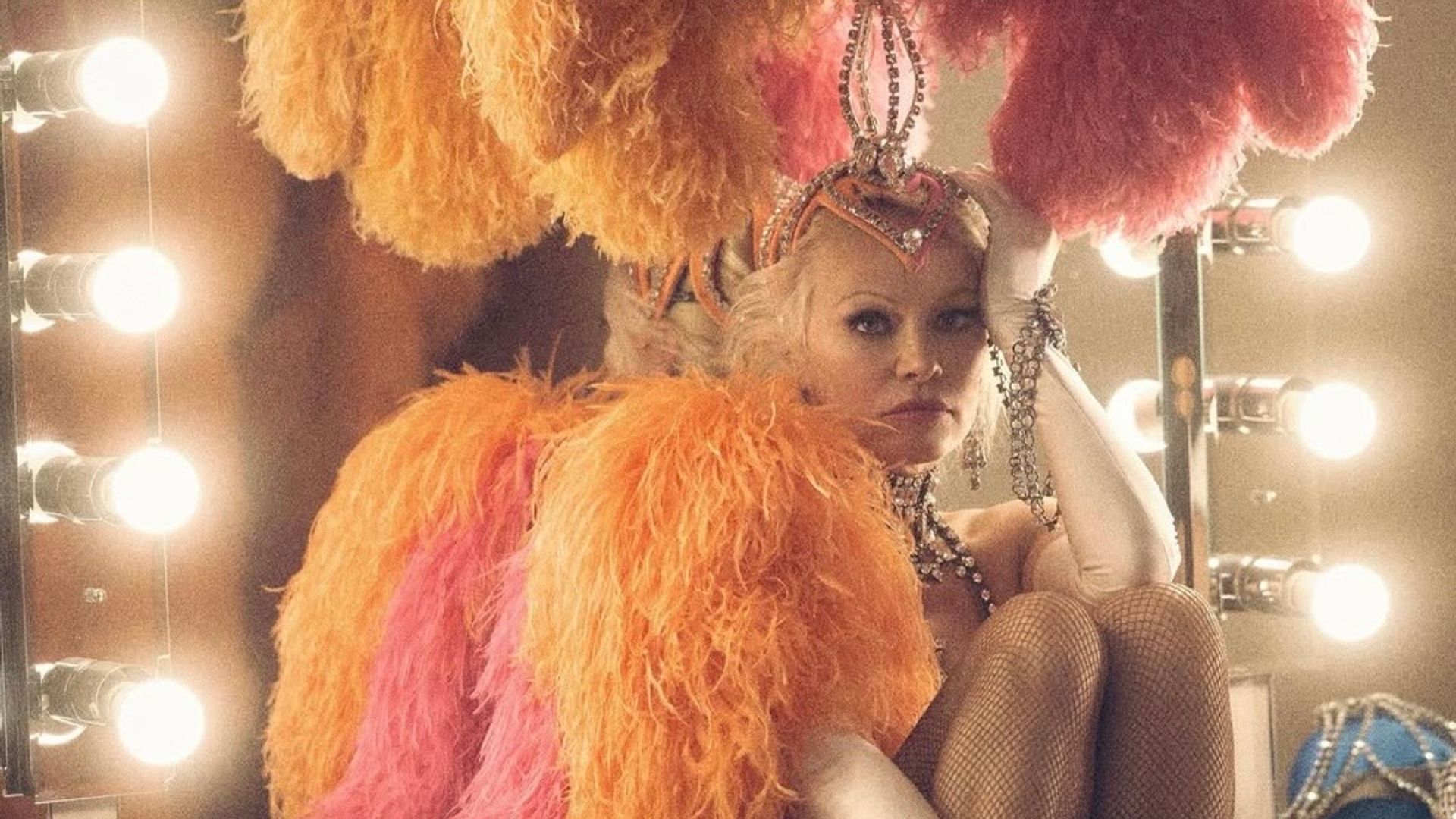 Pamela Anderson in a sparkling show costume with feathers and embellishments sits at a dressing room mirror, thoughtfully resting her head on her hand