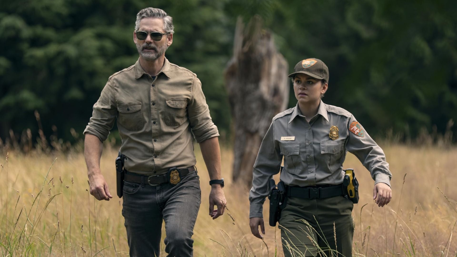 Actor Eric Bana in a green shirt walks with a female police officer across yellow grass with trees in the background