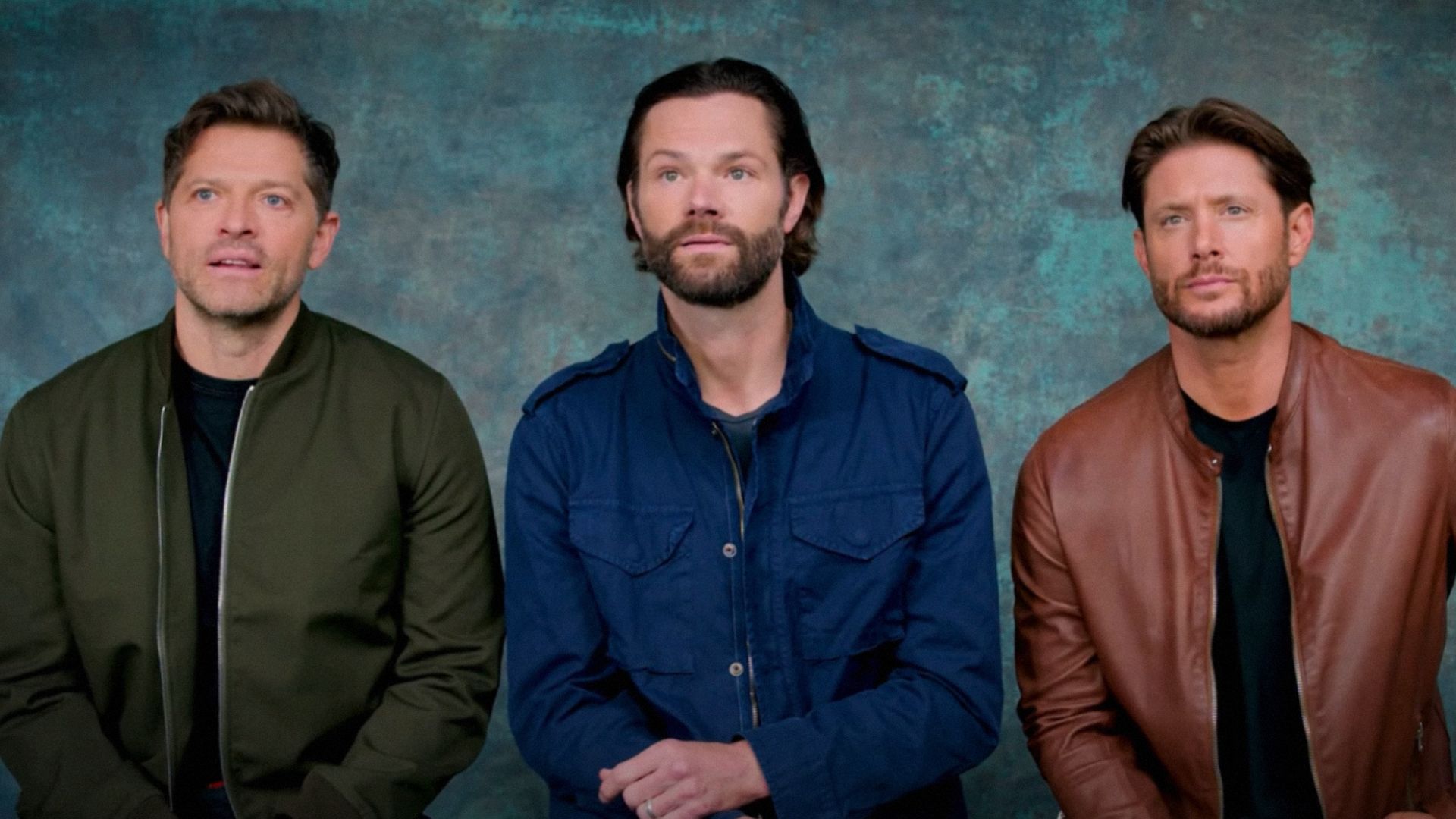 Actors Misha Collins, Jared Padalecki and Jensen Ackles sit against a blue background wearing colorful jackets