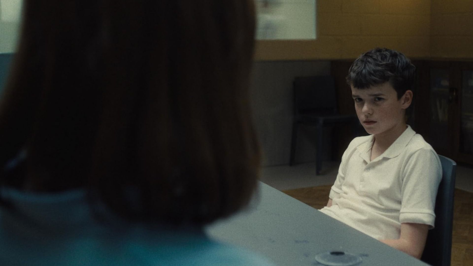 A boy in a white shirt sits at the table and looks at a woman
