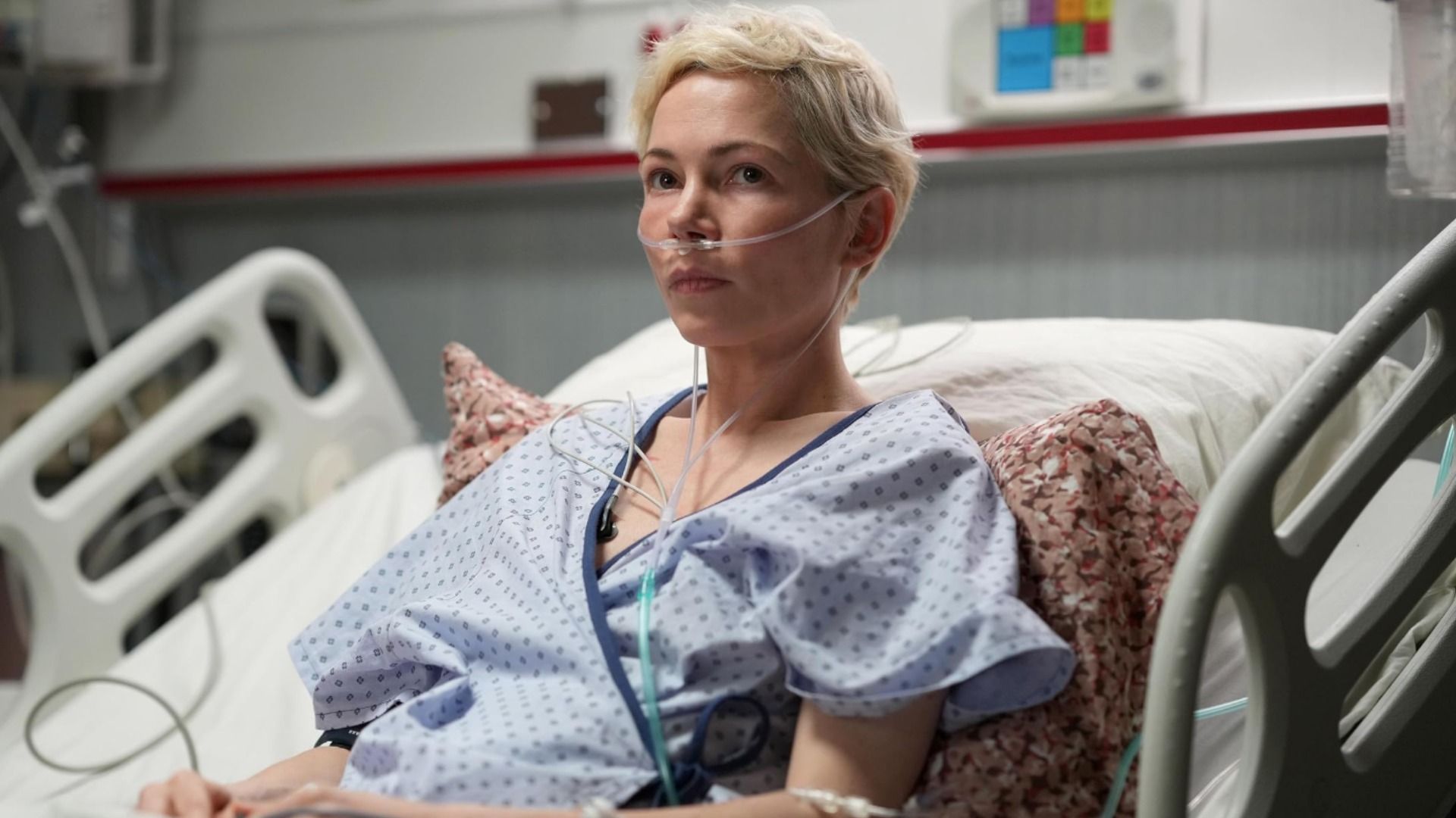 Actress Michelle Williams in hospital patient clothing lies on a bed with a tube in her nose
