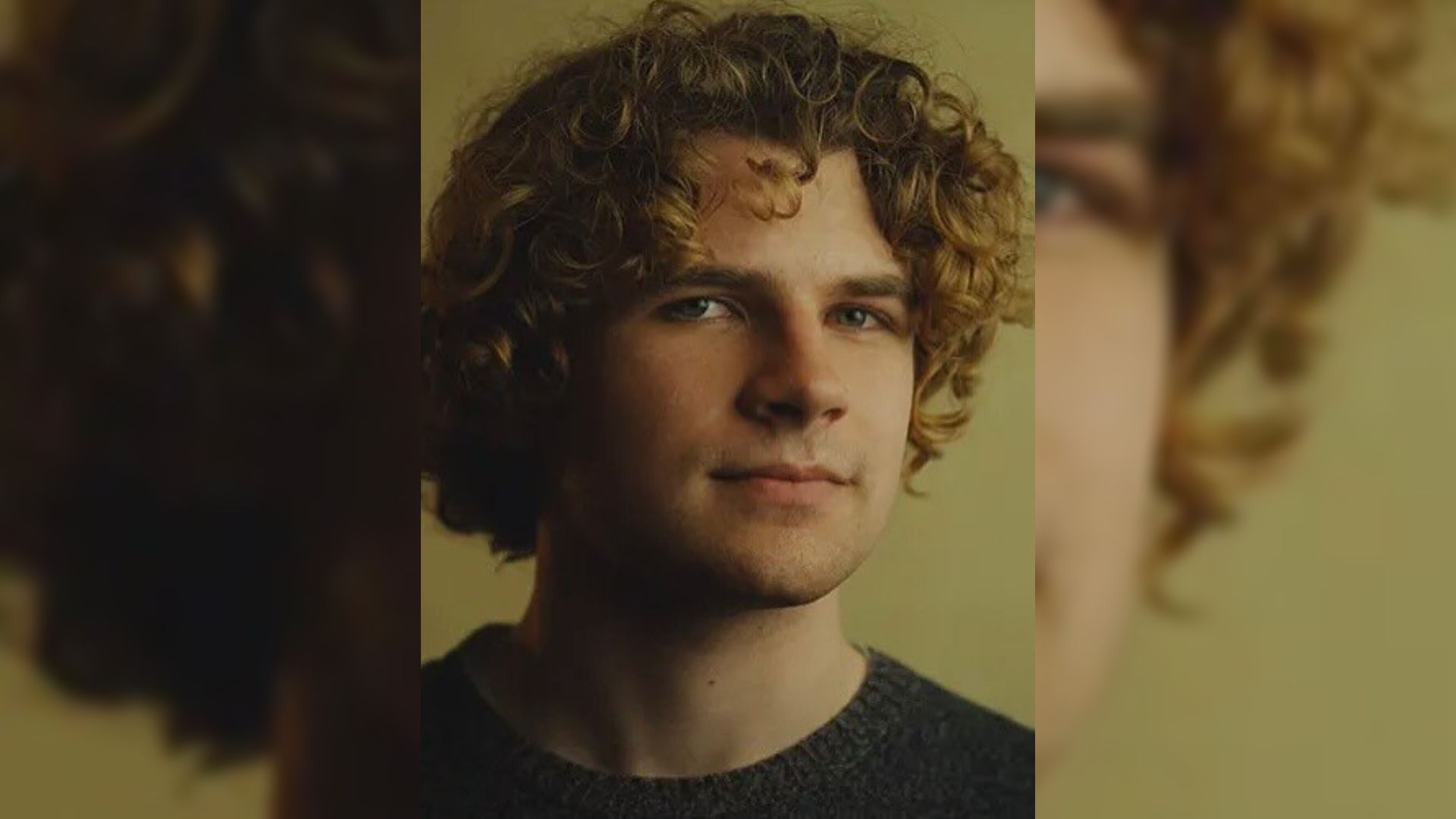A curly-haired young man in a black sweater stands against a yellow background