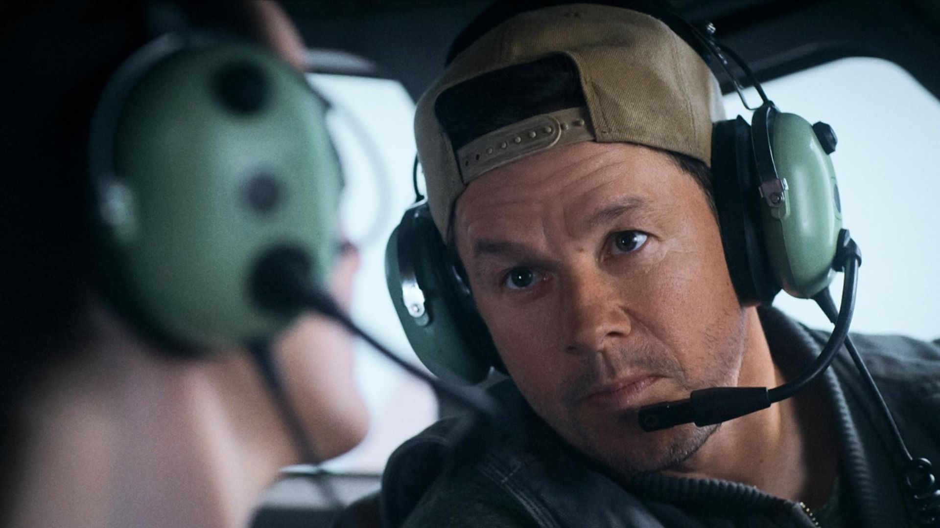 Actor Mark Wahlberg wearing a cap and headphones with microphone sits on an airplane