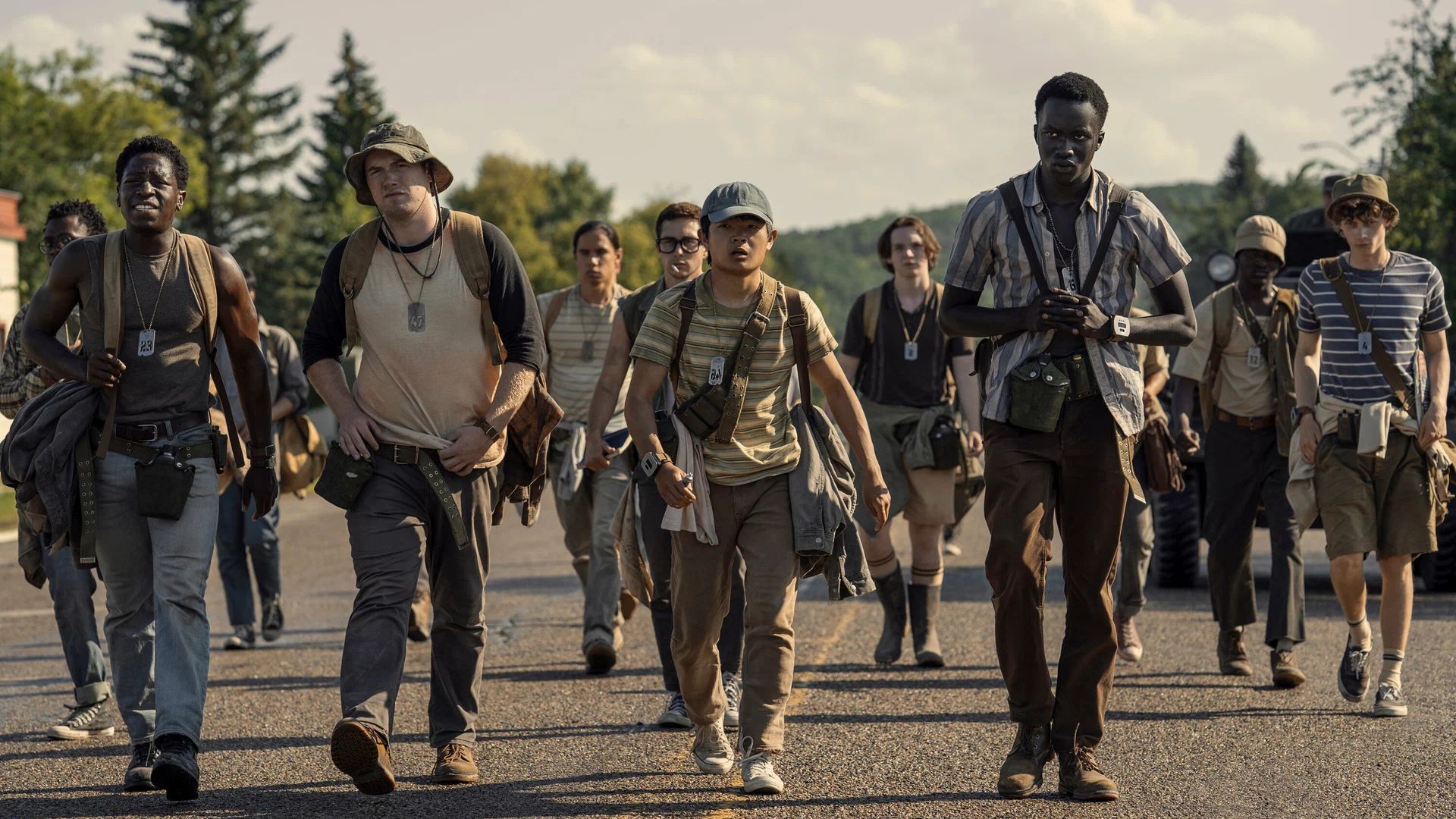 A group of young people in hiking clothes walking along a road on a summer day, with trees and hills in the background