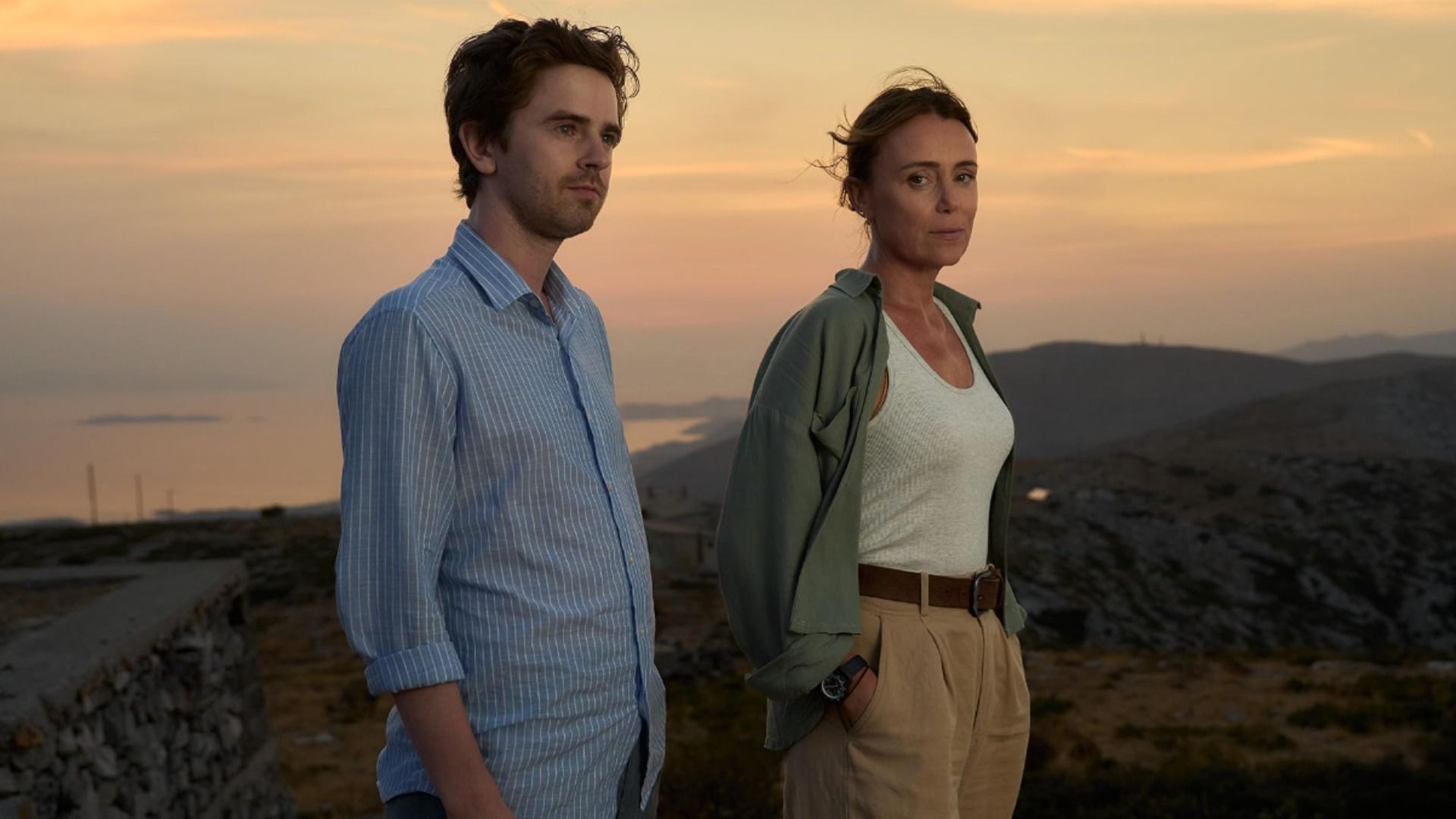 Actor Freddie Highmore in a blue shirt and actress Keeley Hawes in a white tank top standing against a mountain backdrop