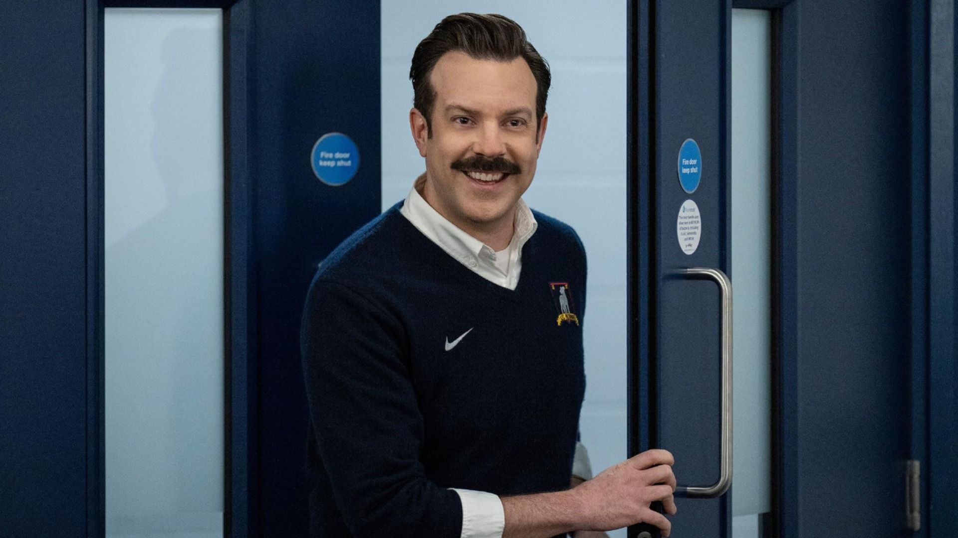 Actor Jason Sudeikis in a blue sweater and with a mustache stands near a door and smiles