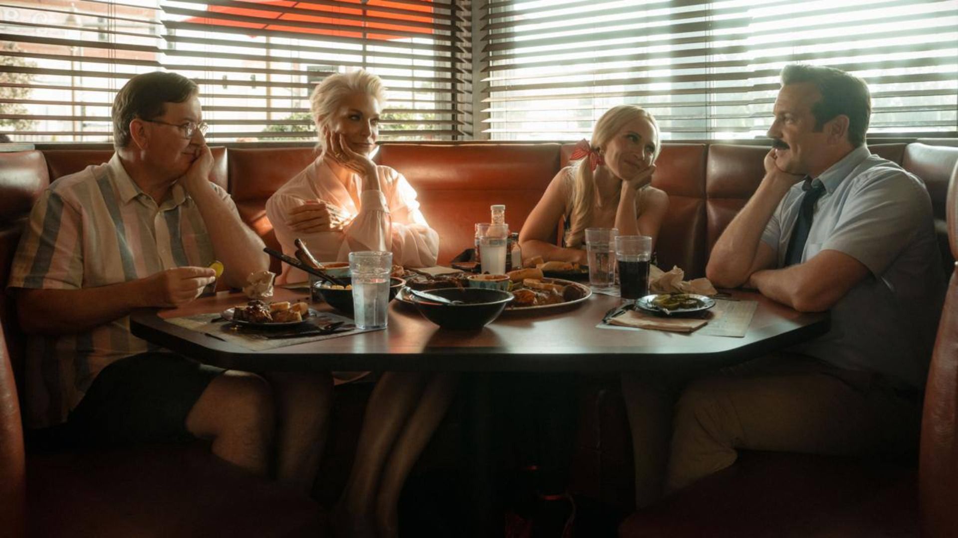 Actor Jason Sudeikis with a mustache sits at a set table surrounded by people