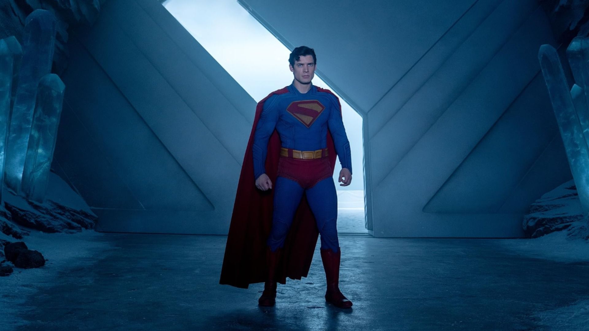 Actor David Corenswet in a blue and red superhero costume stands by large gates