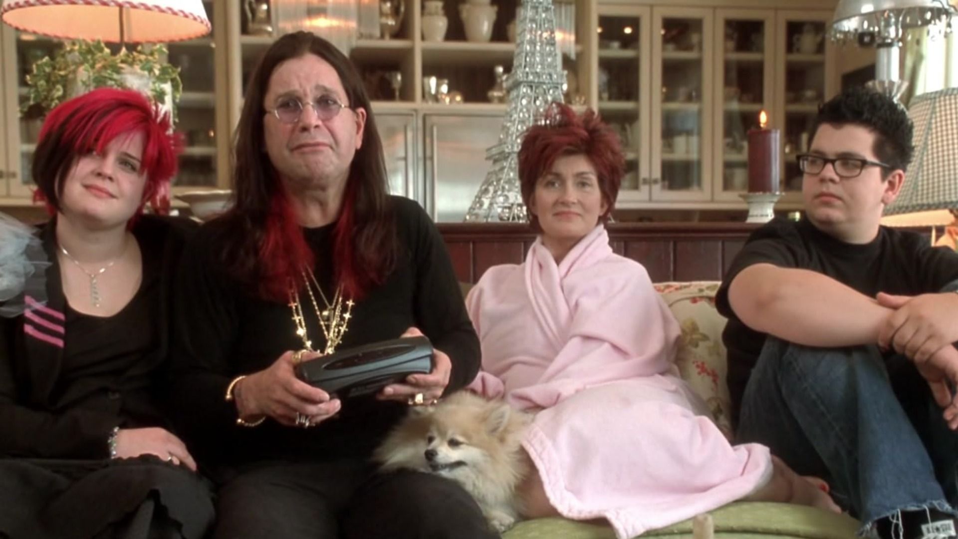 Singer Ozzy Osbourne in a black suit with chains sits on a couch with his wife and children