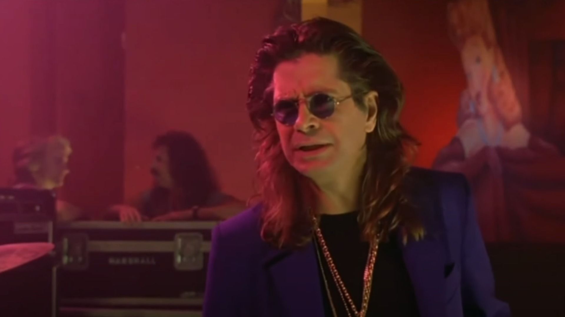 Singer Ozzy Osbourne in a blue blazer and gold chain stands on stage