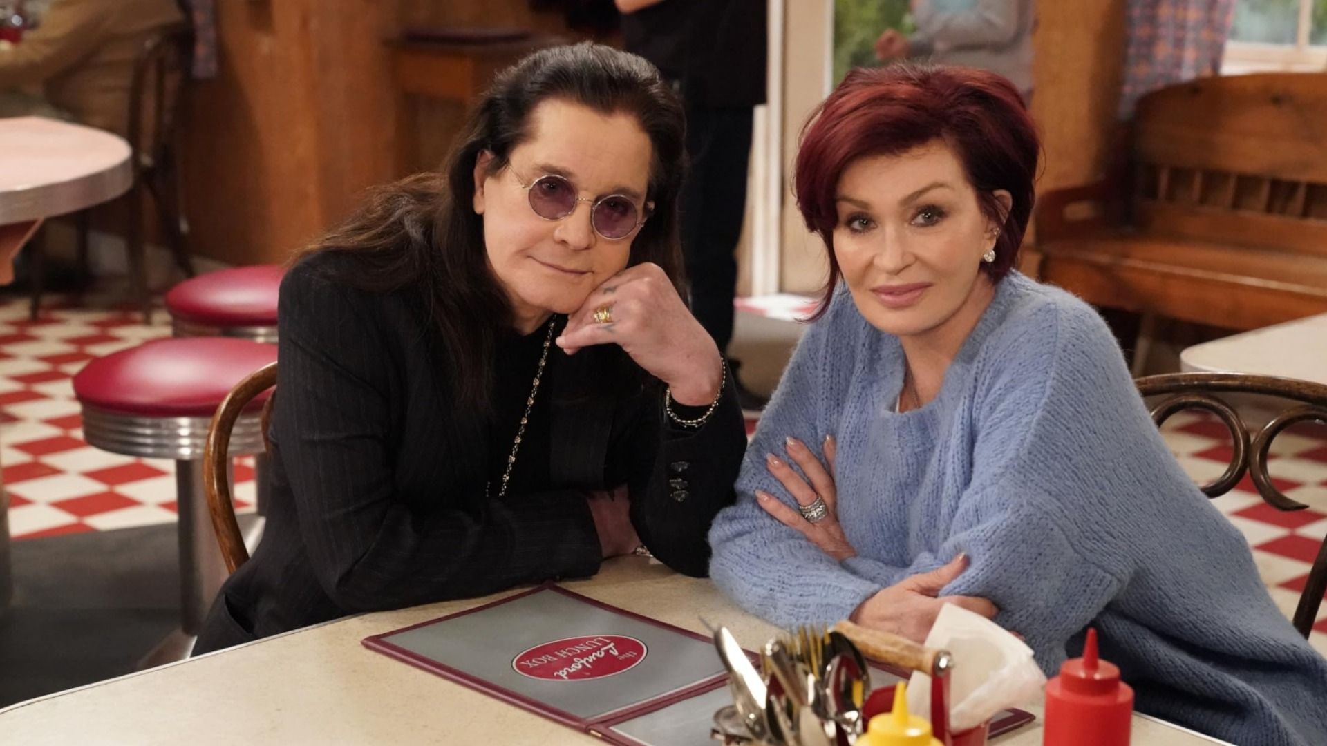 Singer Ozzy Osbourne wearing glasses and a black jacket sits at a table with his wife