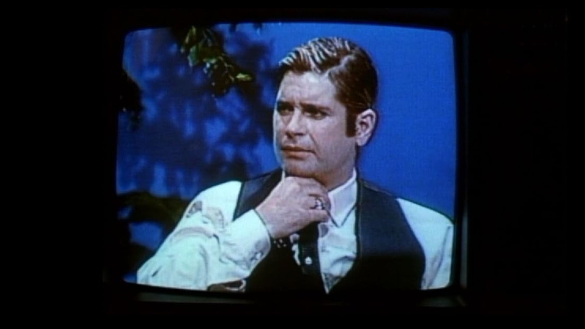 Singer Ozzy Osbourne with short hair sits at a table wearing a white shirt and black vest