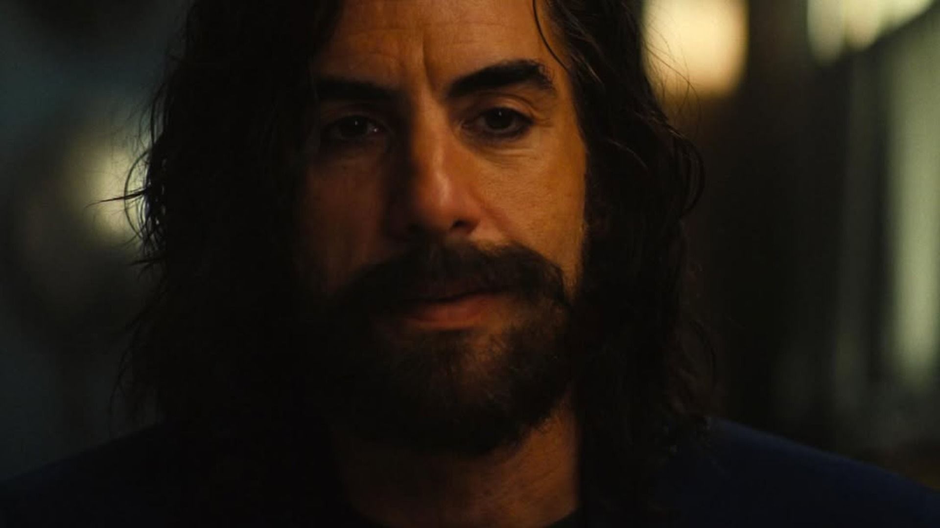 Actor Sacha Baron Cohen with a beard and long hair stands against a blurred background