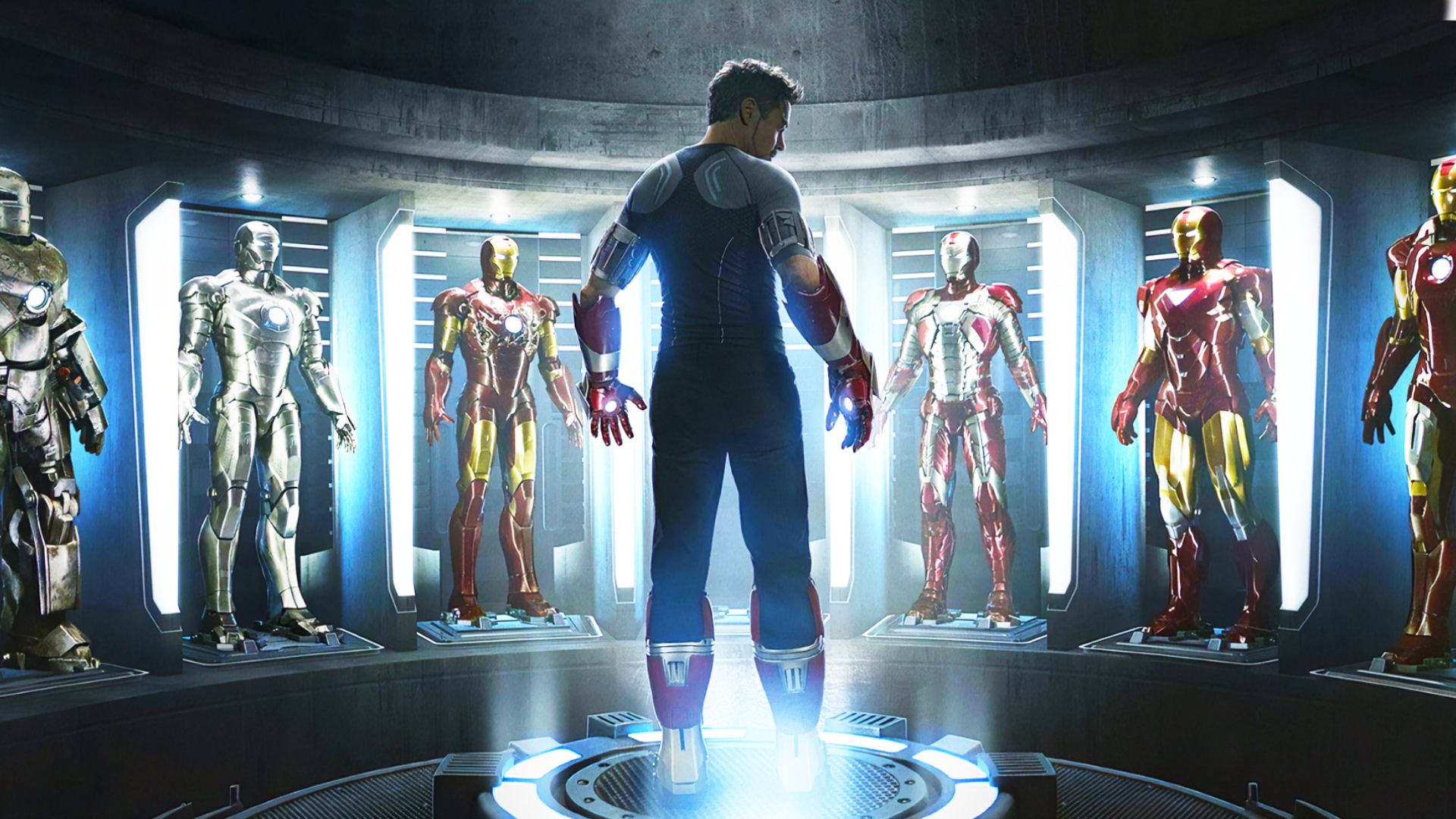 Actor Robert Downey Jr. in gray and black clothing stands surrounded by Iron Man suits
