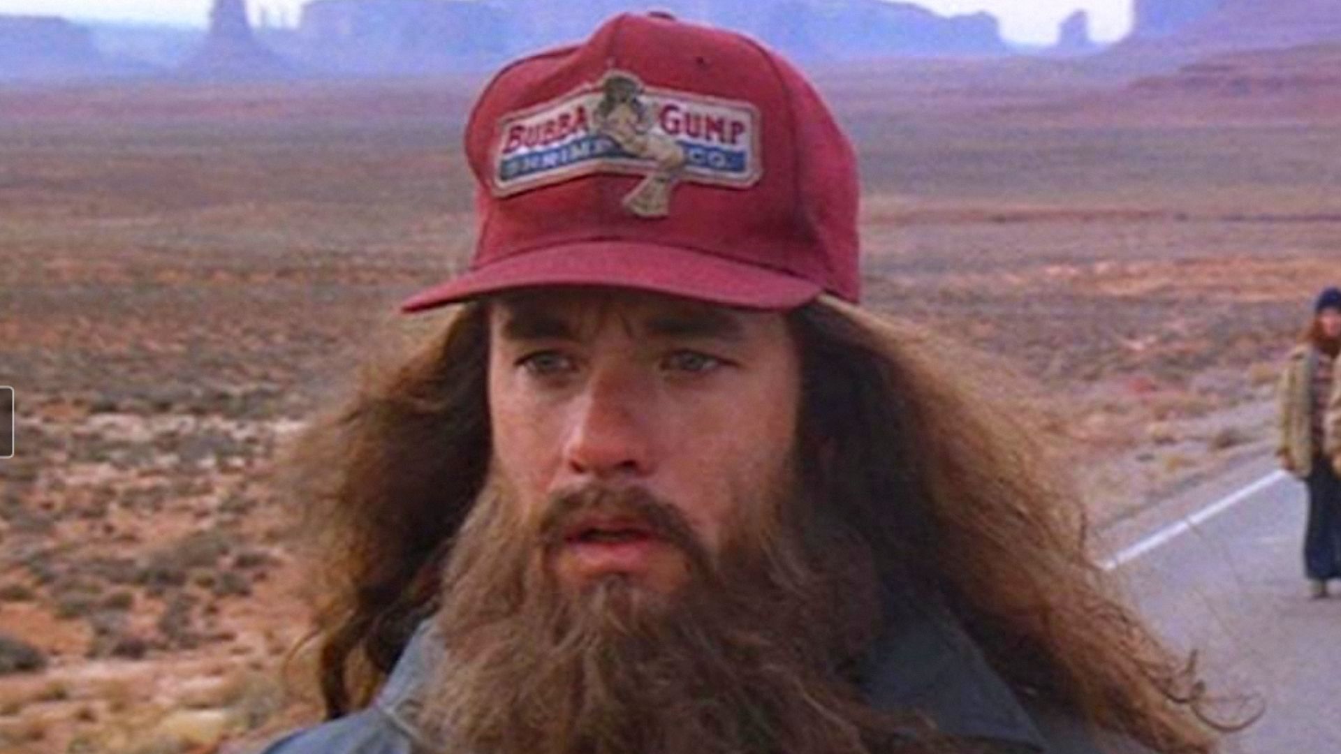 Actor Tom Hanks with long hair and a beard stands wearing a red cap