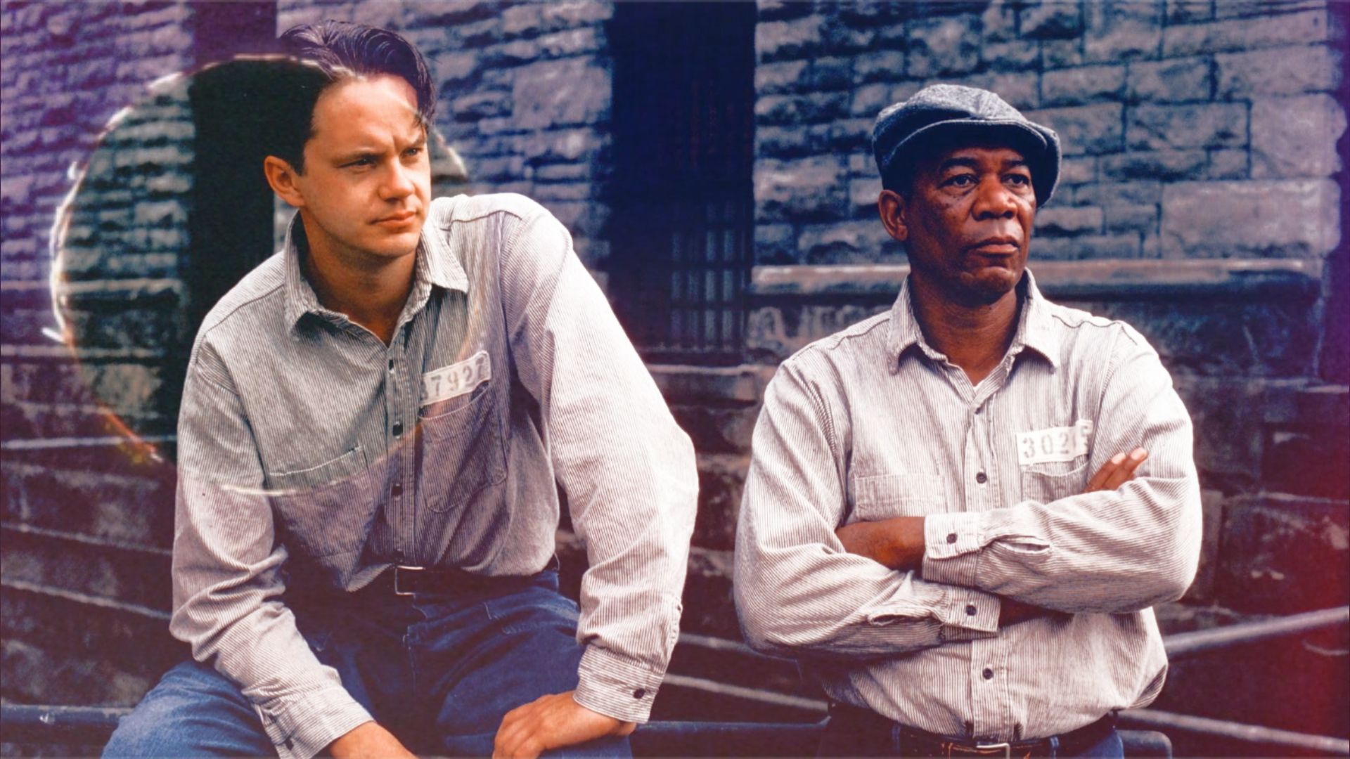 Actors Tim Robbins and Morgan Freeman in prison uniforms sitting by a stone wall