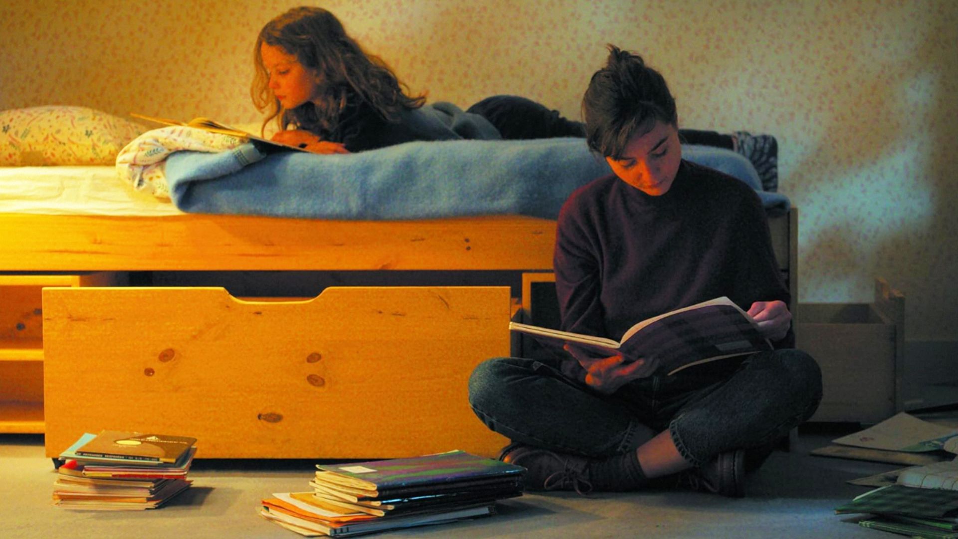 A girl lies on the bed reading a book while a woman sits beside the bed with a book