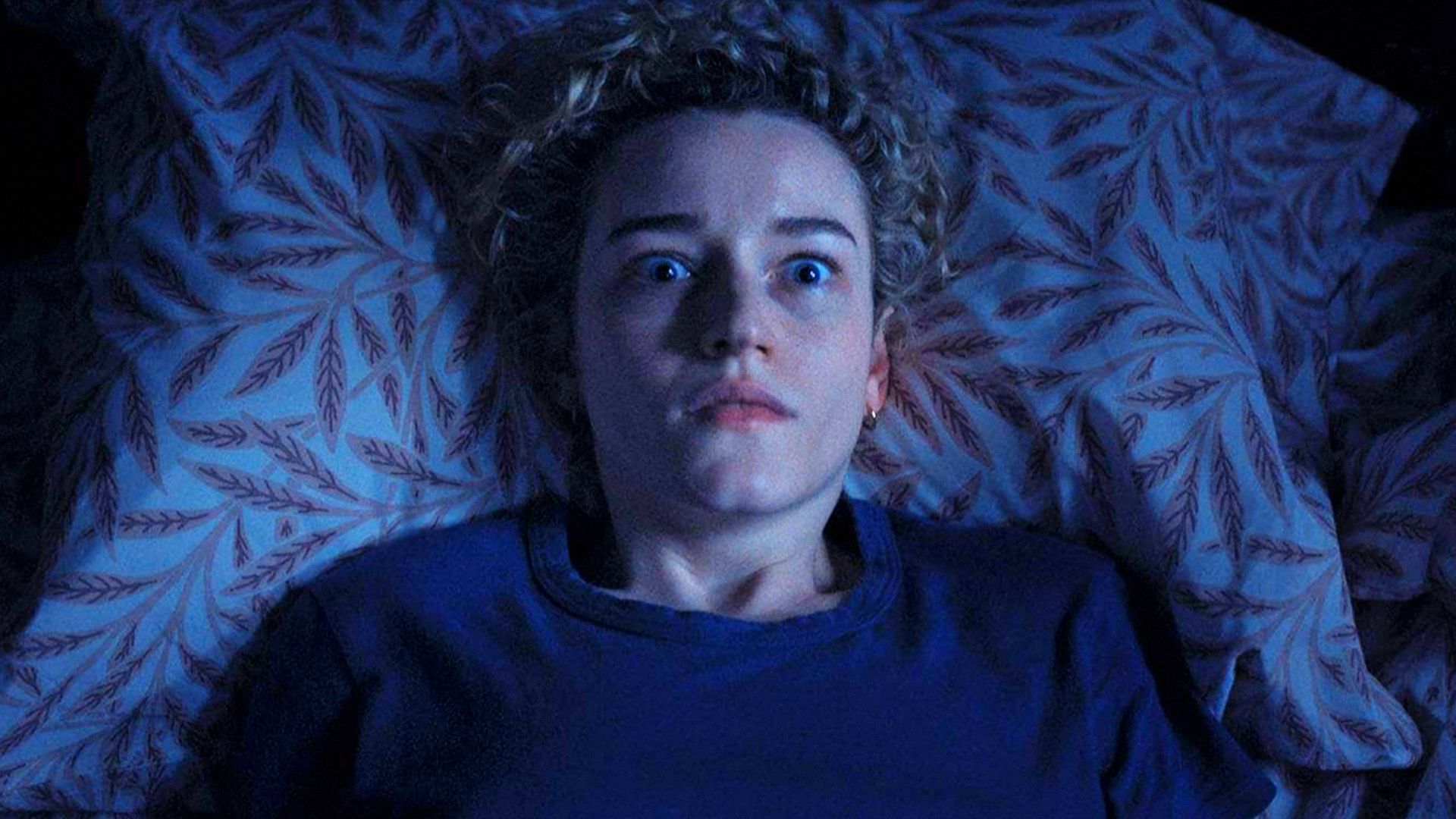 Actress Julia Garner in a blue sweater lies on a bed, frightened by something