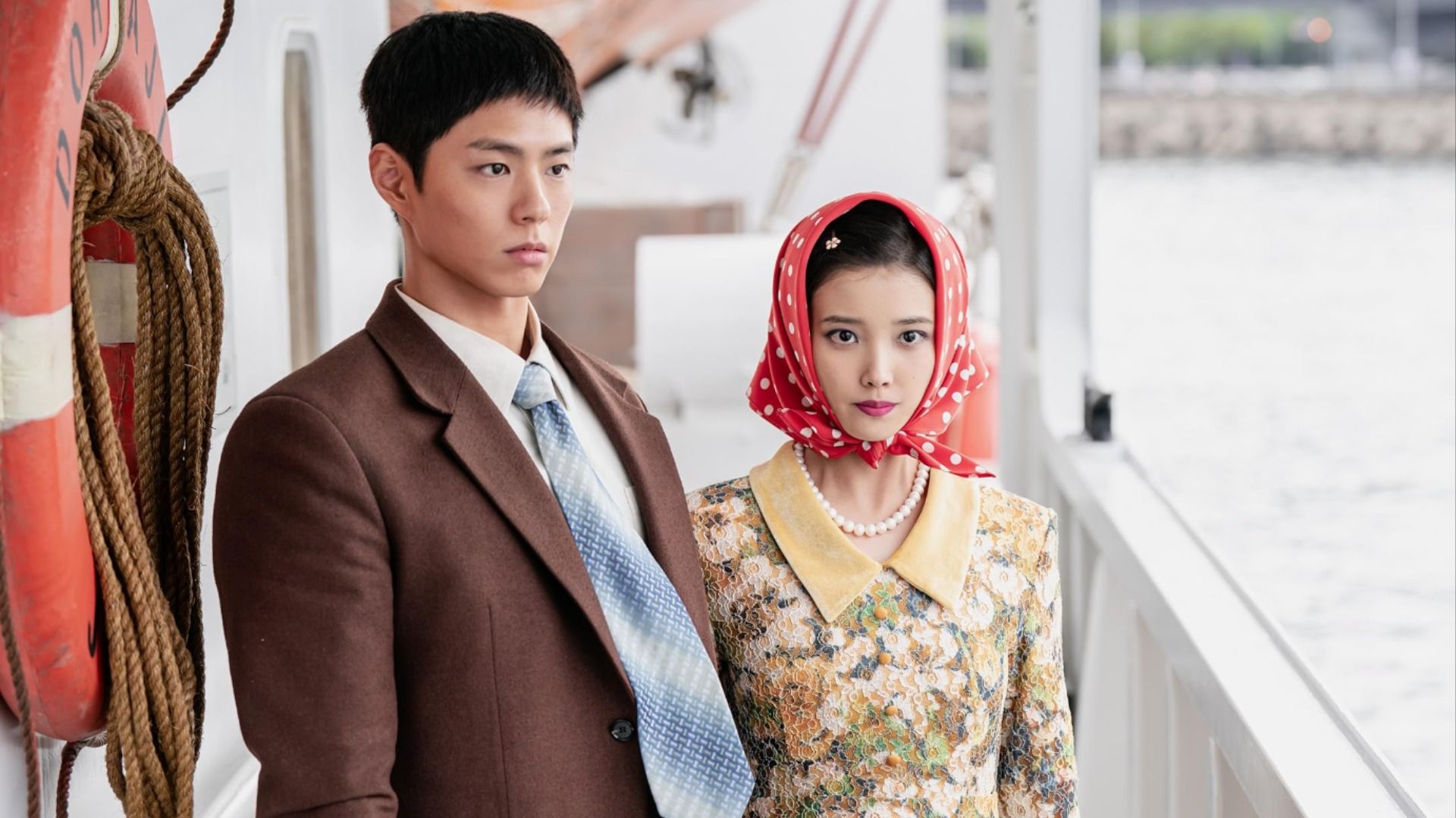 A guy in a brown blazer with a tie stands next to a girl in a yellow-green dress with a red headscarf