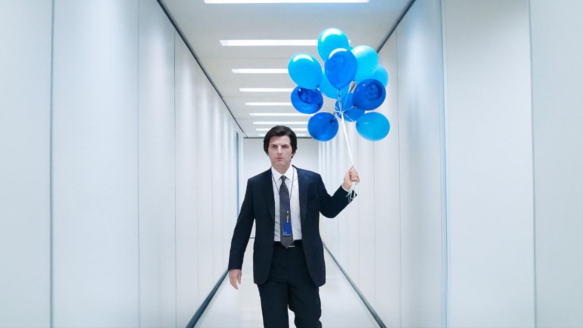 Actor Adam Scott in a classic suit walks down a white hallway holding blue balloons