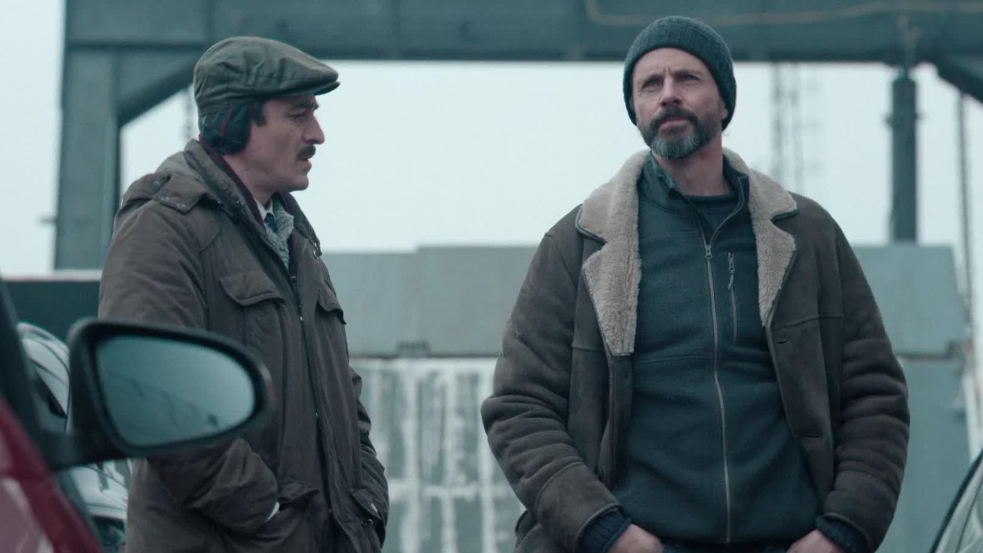 Two men in gray jackets standing next to a red car