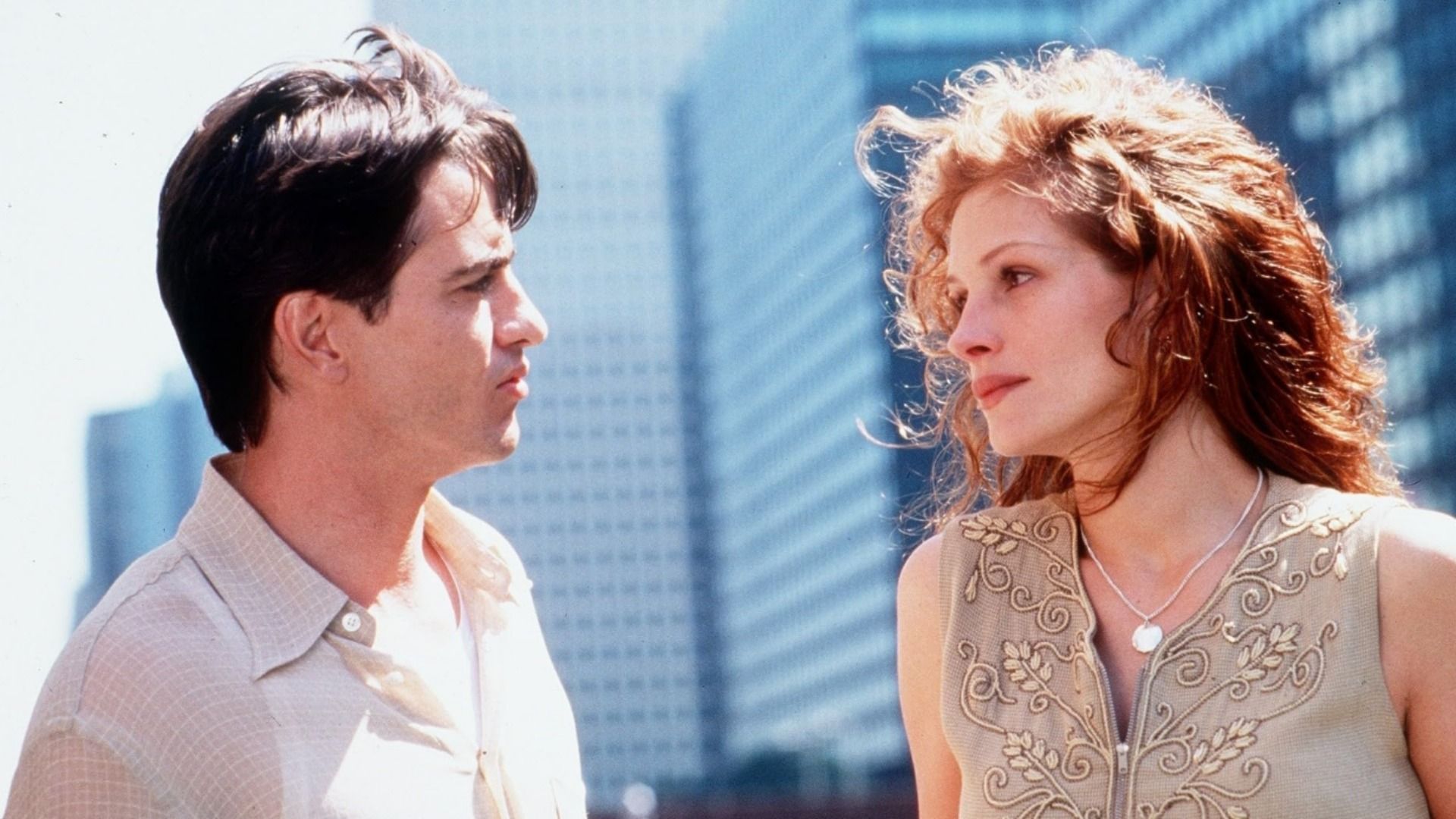 Actor Dermot Mulroney in a light shirt looks at actress Julia Roberts against a backdrop of high-rise buildings