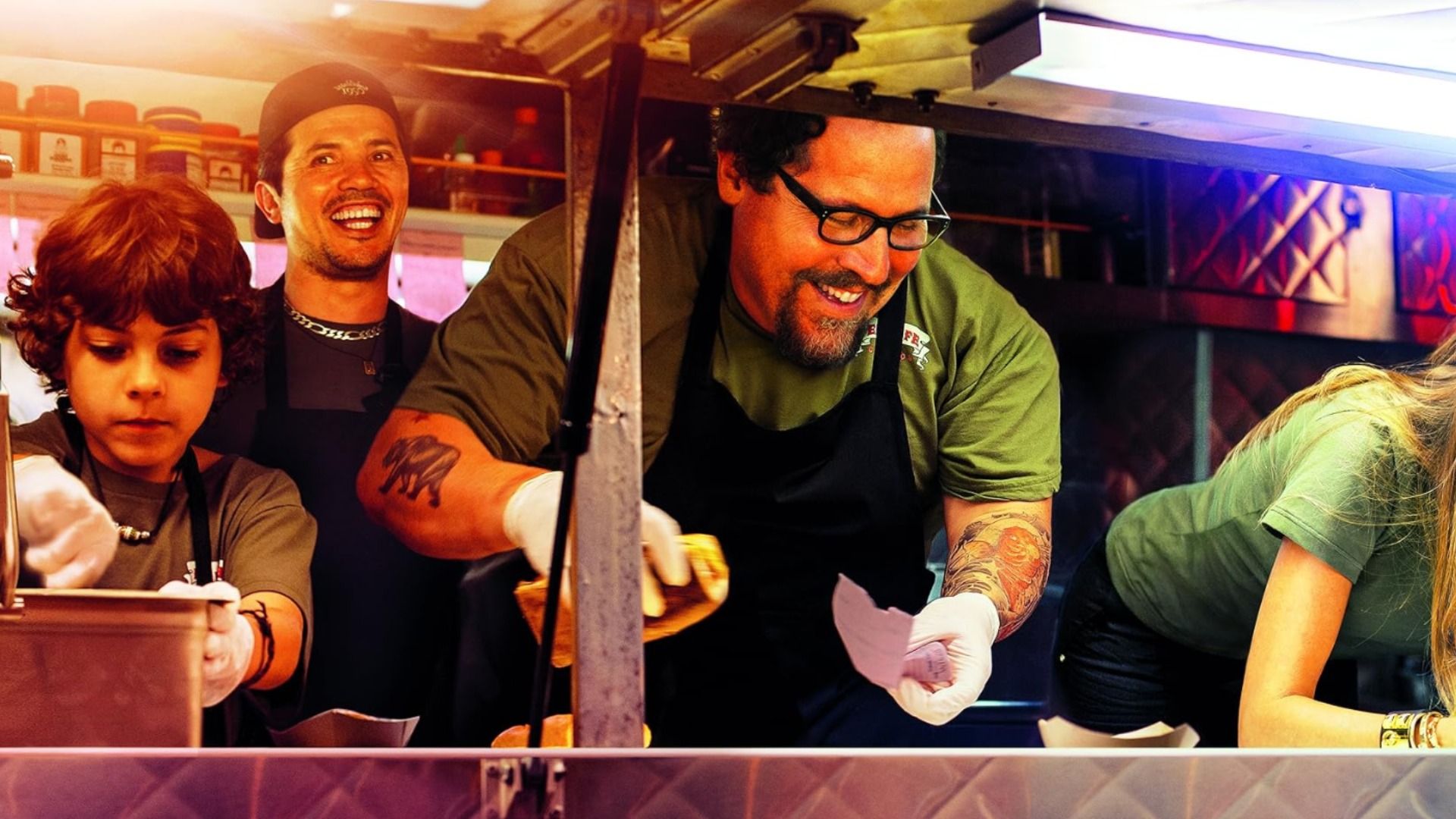 Actor Jon Favreau in a black apron holds a bun while surrounded by a child, a man and a woman