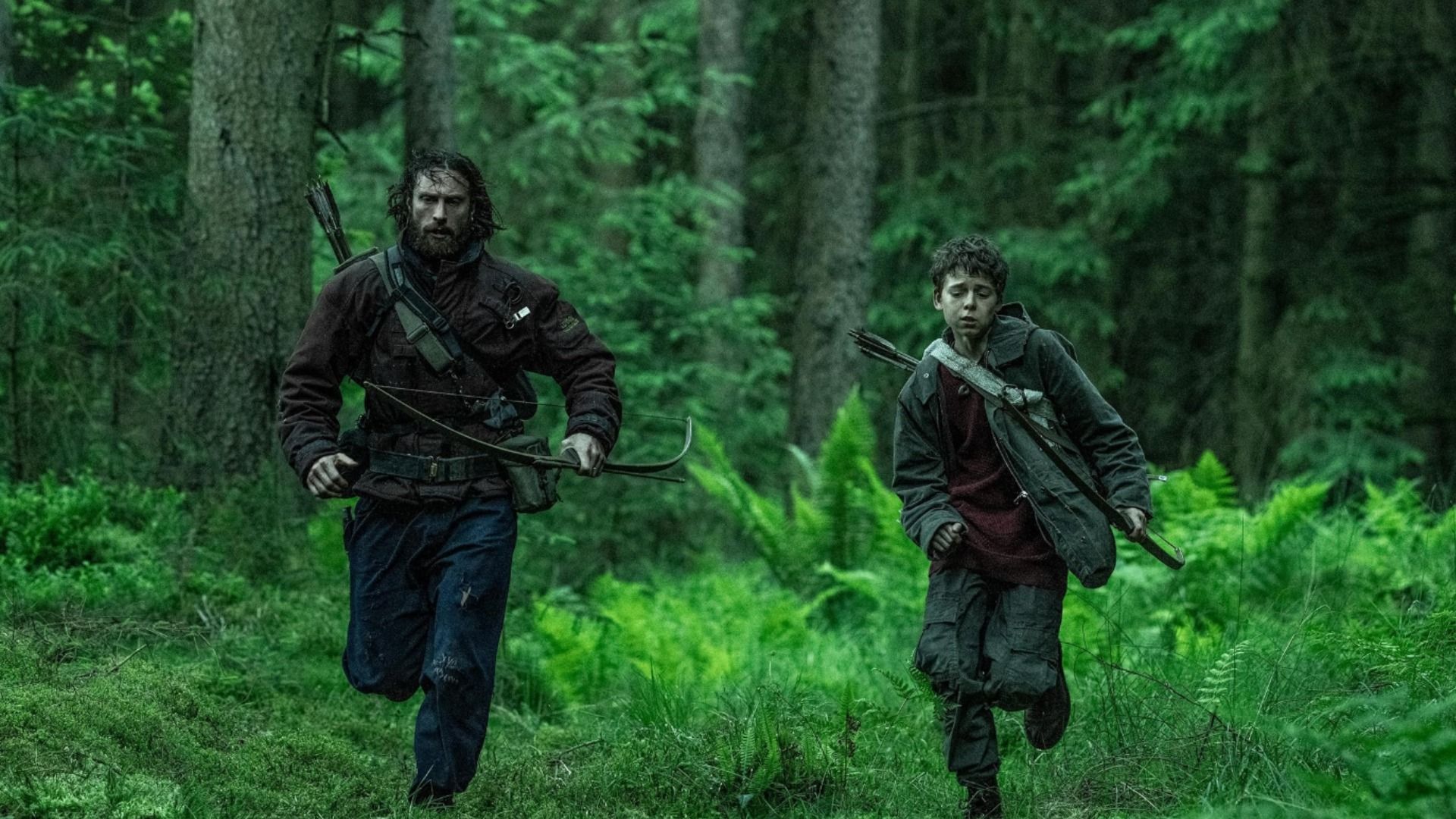 A man and child in dark clothing run through the forest with bows and arrows in their hands