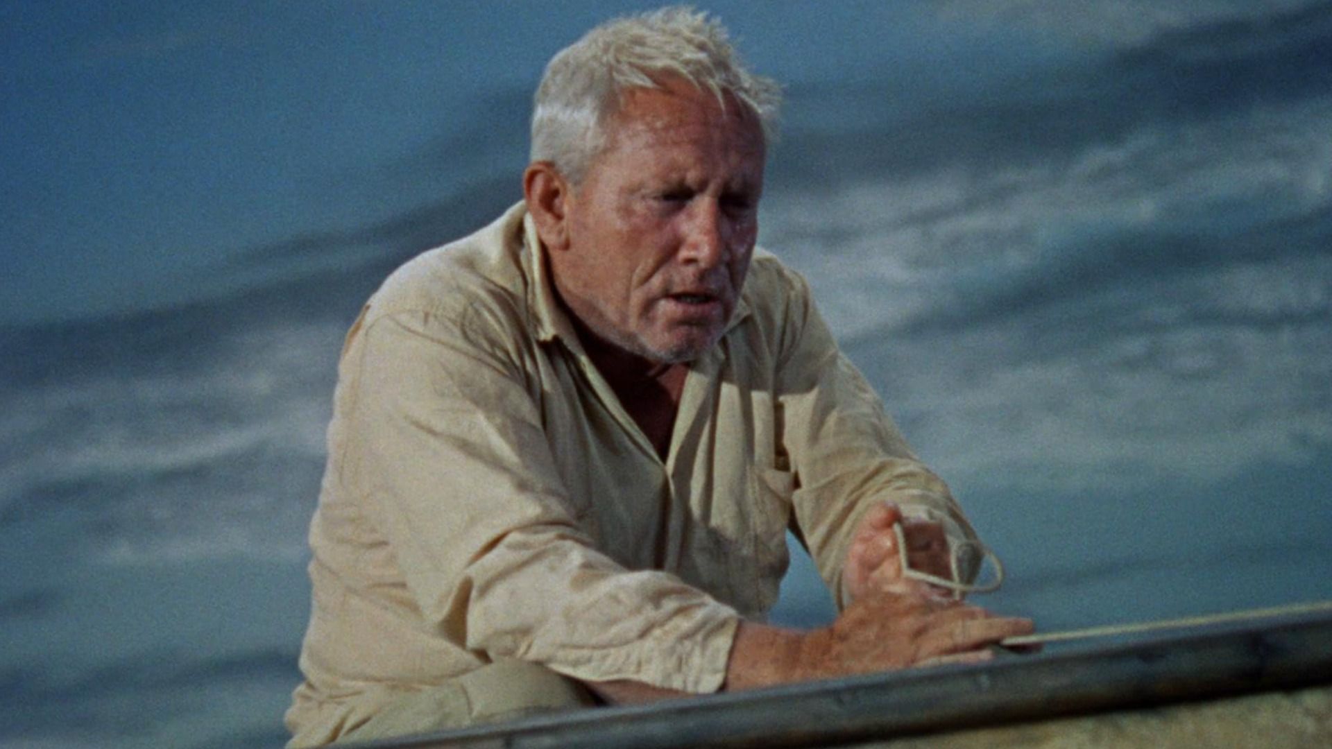 A man in a white shirt with white hair stands by a boat looking off to the side