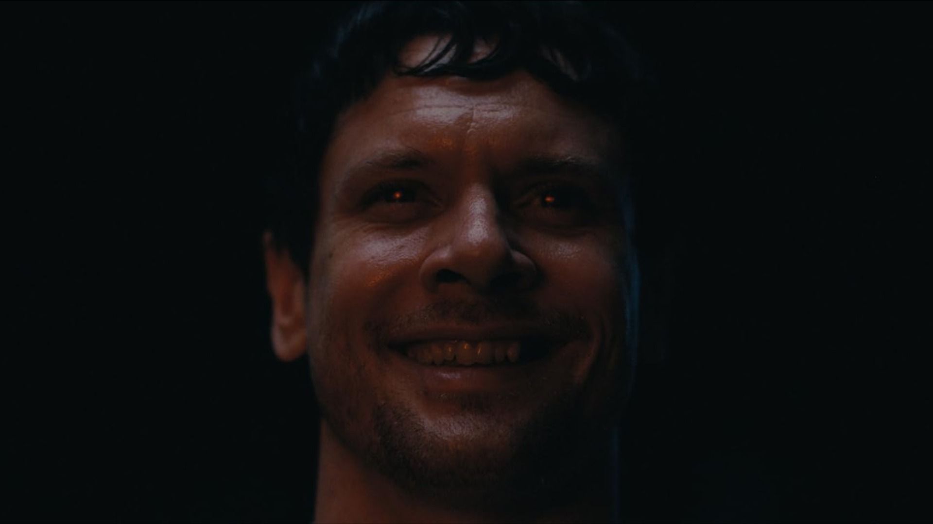 A man smiles against a black background with a red gleam in his eyes