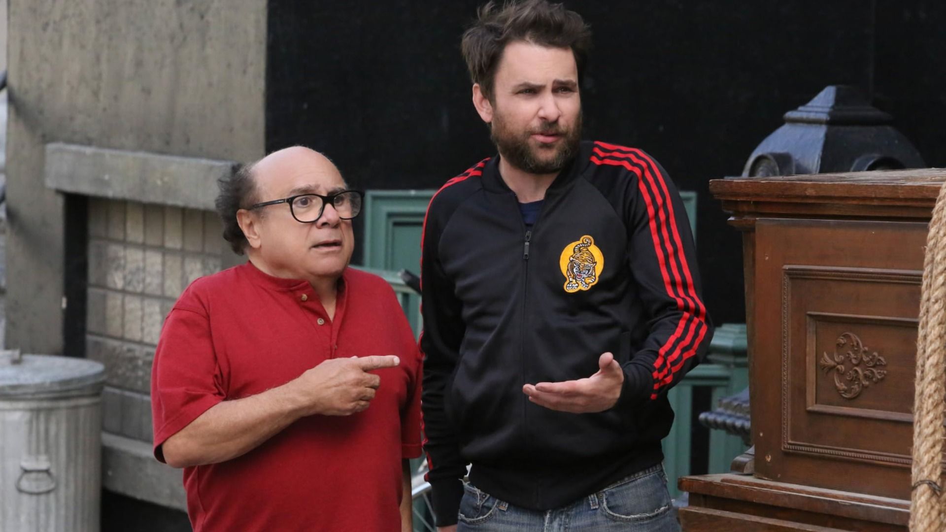 Actor Danny DeVito in a red shirt points his finger at a man in athletic wear