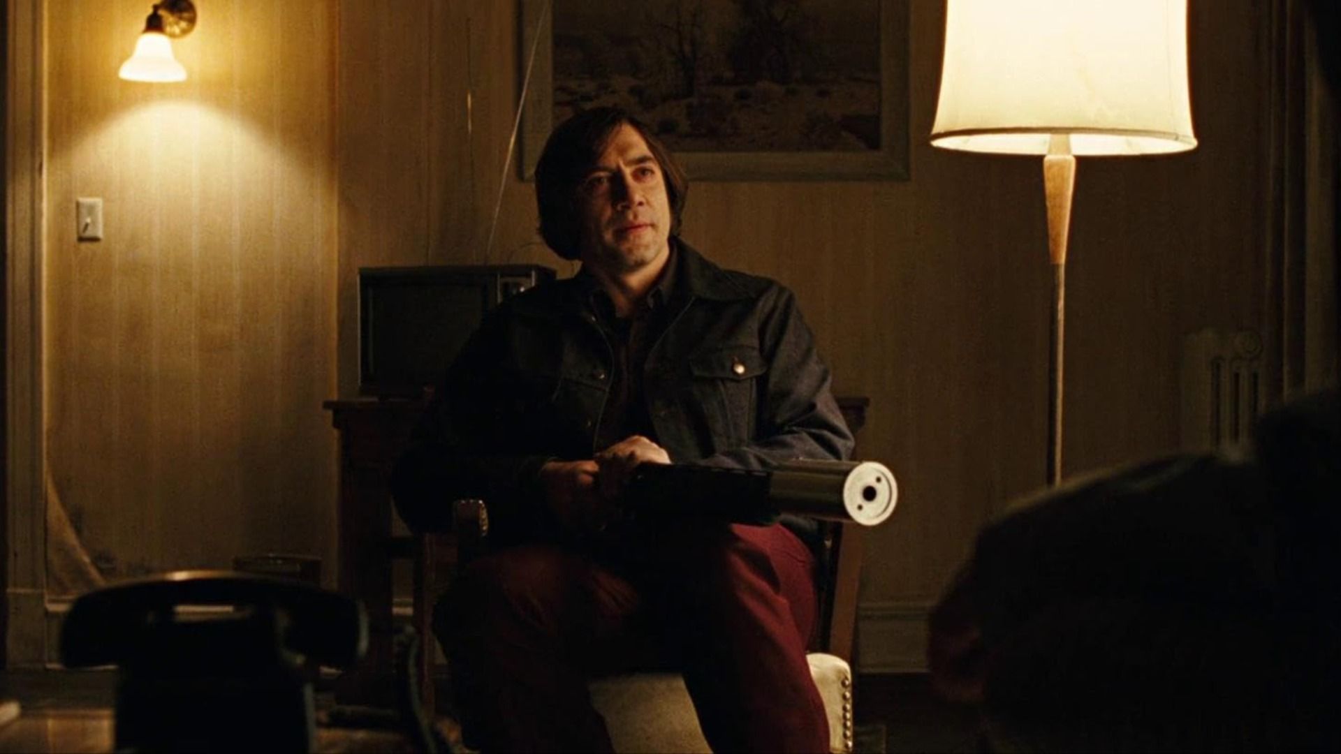 Actor Javier Bardem with long hair sits in a chair holding a weapon